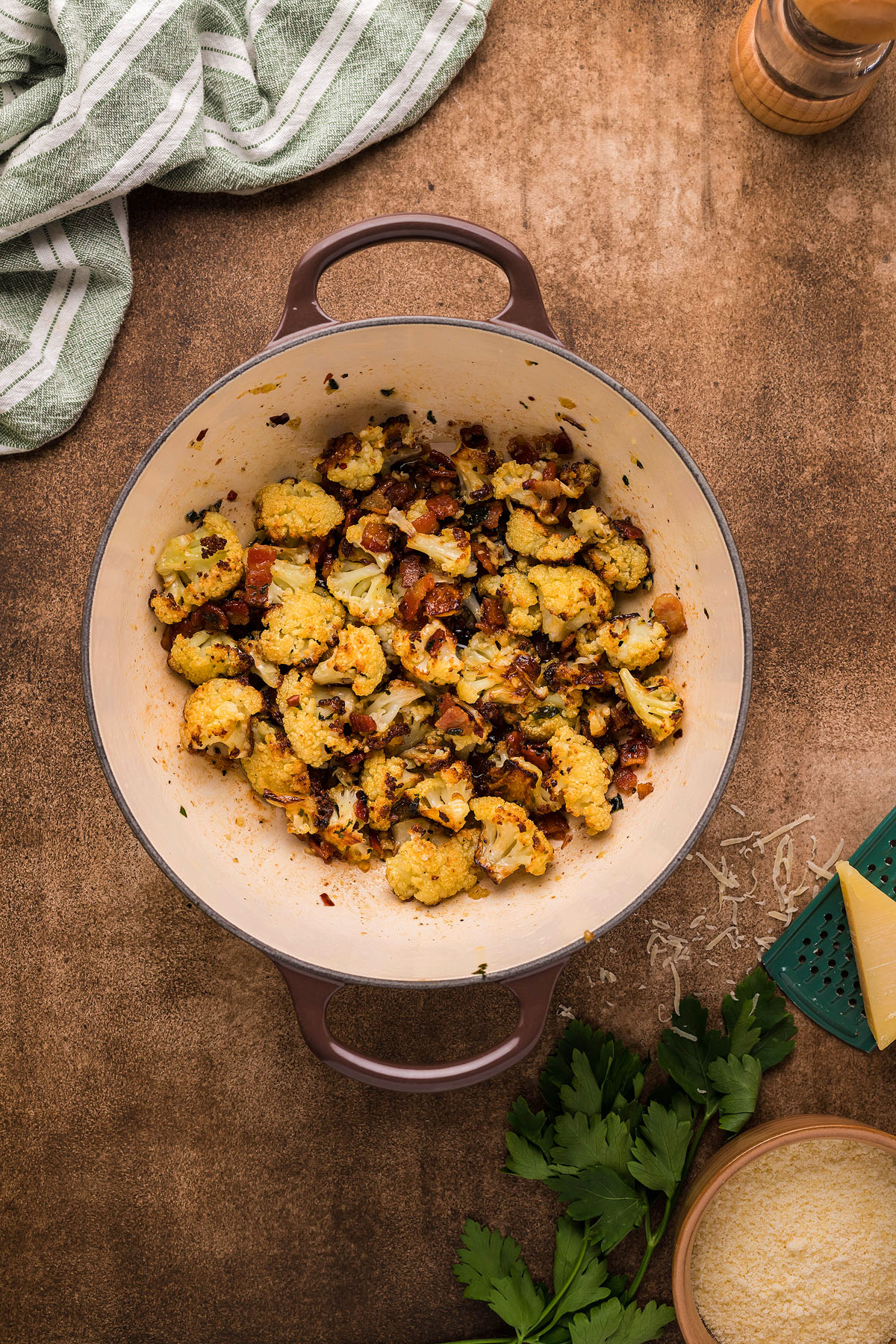 Cooked cauliflower with herbs and bacon in a large pot, perfect for an easy weeknight dinner inspired by Roasted Cauliflower Carbonara, surrounded by cheese, parsley, and a green towel.