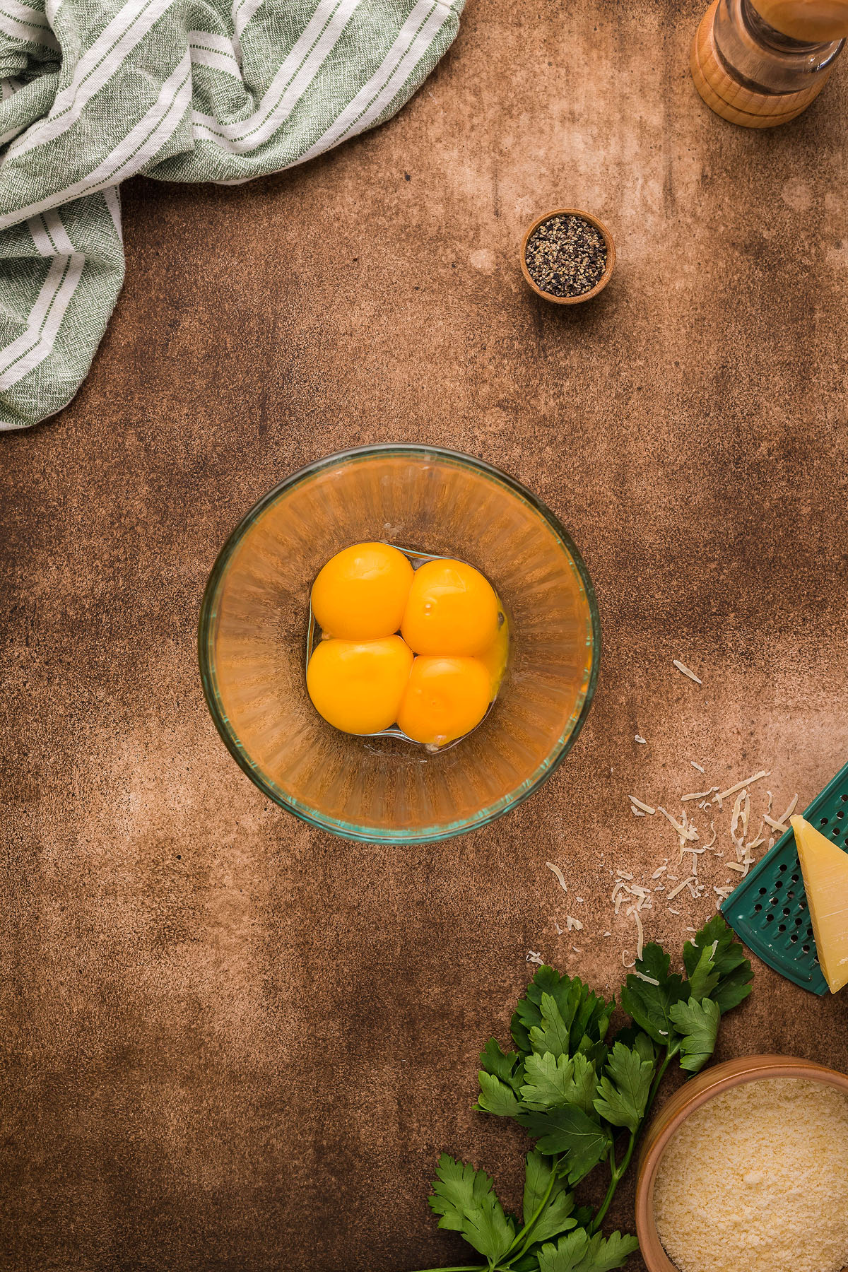 Four egg yolks in a glass bowl on a brown surface, surrounded by herbs, cheese, pepper, and a striped towel—perfect for crafting an easy weeknight dinner like Roasted Cauliflower Carbonara with creamy cauliflower sauce.