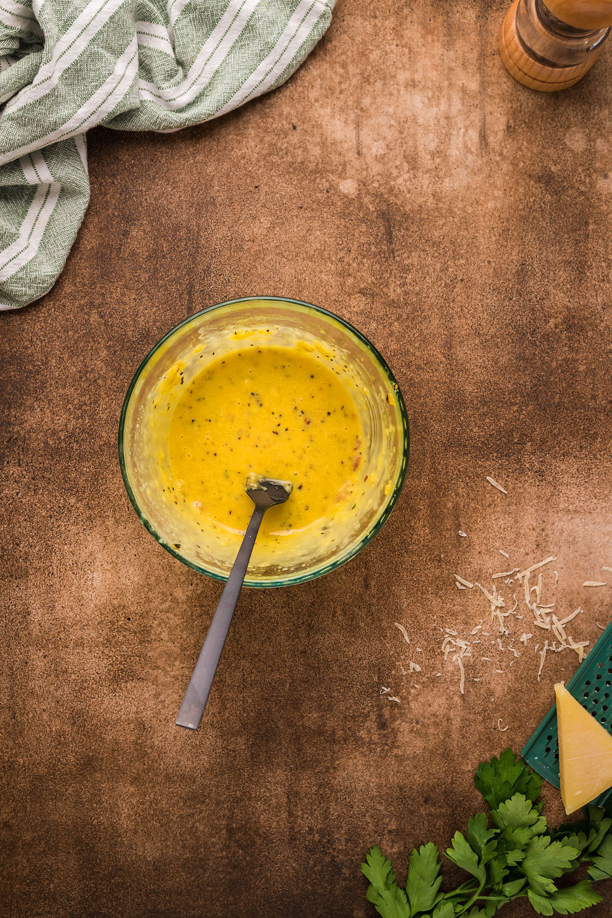 A bowl of creamy cauliflower sauce with a spoon sits on a brown surface, next to grated cheese, parsley, and a striped towel—perfect for Roasted Cauliflower Carbonara and an easy weeknight dinner.