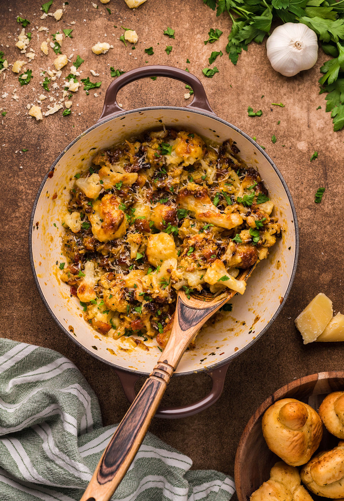 Cheesy baked cauliflower casserole in a pot with a wooden spoon, surrounded by herbs and garlic—an easy weeknight dinner inspired by Roasted Cauliflower Carbonara and rich cauliflower sauce.