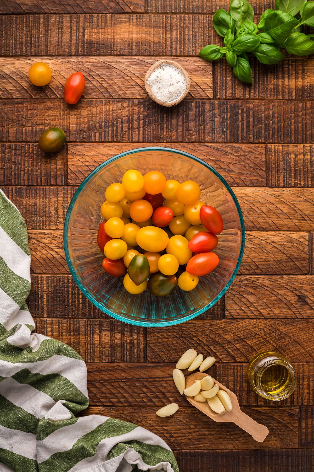 A glass bowl of colorful cherry tomatoes on a wooden surface with garlic, olive oil, salt, basil, and a towel nearby—perfect ingredients for Italian meatball dinner ideas or a gluten free meatball recipe.