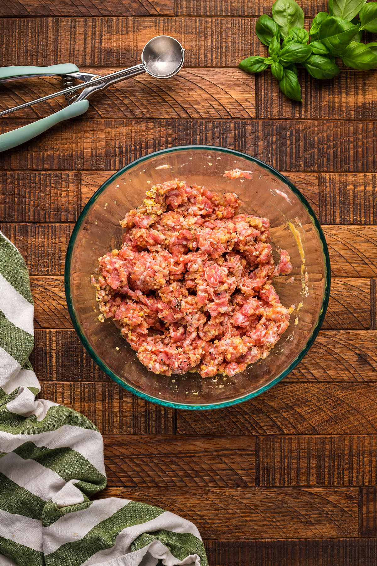 A glass bowl of raw ground meat mixture on a wooden surface, perfect for crafting a gluten free meatball recipe, with herbs, a scoop, and a striped cloth nearby.