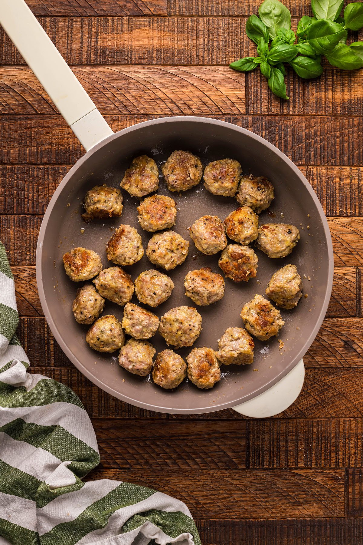 Meatballs cooking in a frying pan on a wooden surface, with fresh basil and a striped towel nearby—perfect for gluten free meatball recipe inspiration or Italian meatball dinner ideas.