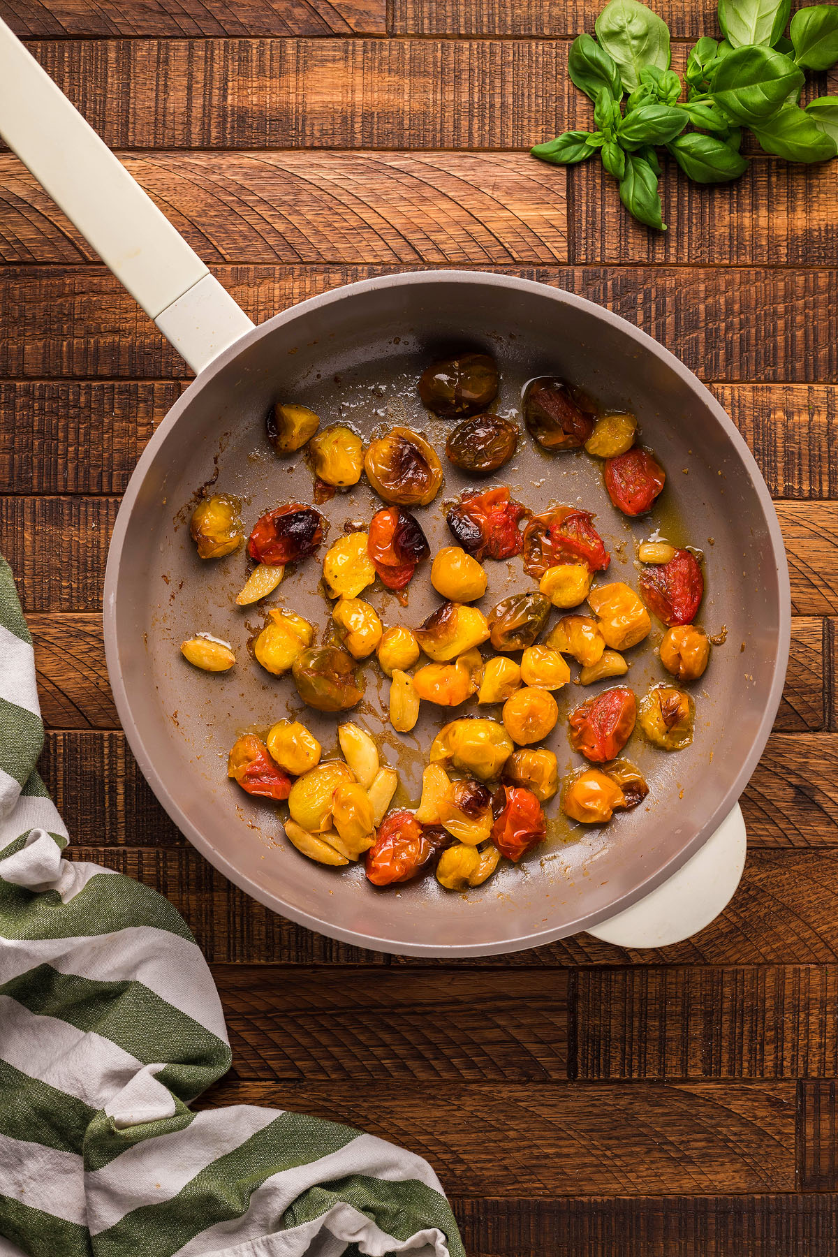 Sautéed cherry tomatoes in a frying pan on a wooden surface, with basil and a striped towel nearby—perfect as a topping for roasted tomato and meatball polenta or served alongside your favorite gluten free meatball recipe.