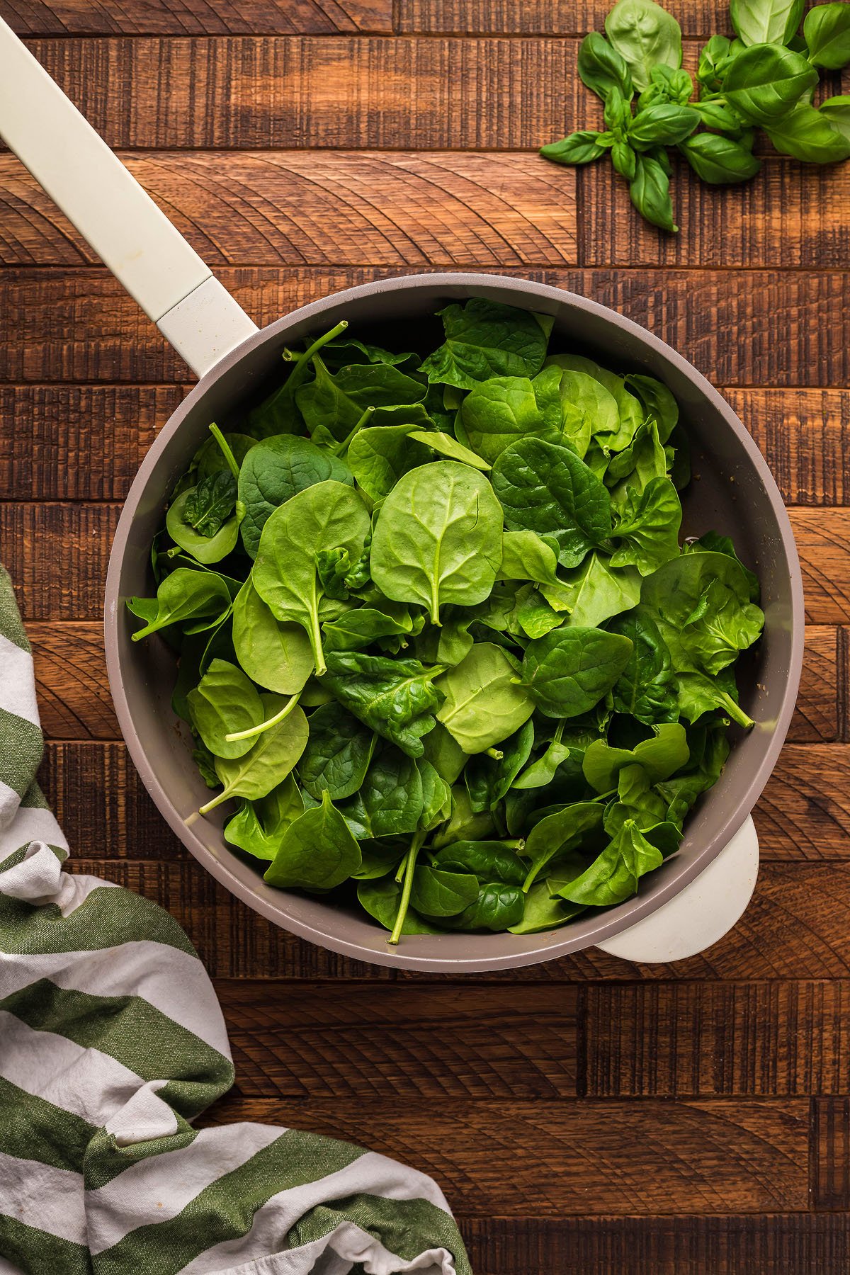 A pan filled with fresh spinach leaves on a wooden surface, perfect for pairing with your favorite gluten free meatball recipe, with basil and a striped towel nearby.
