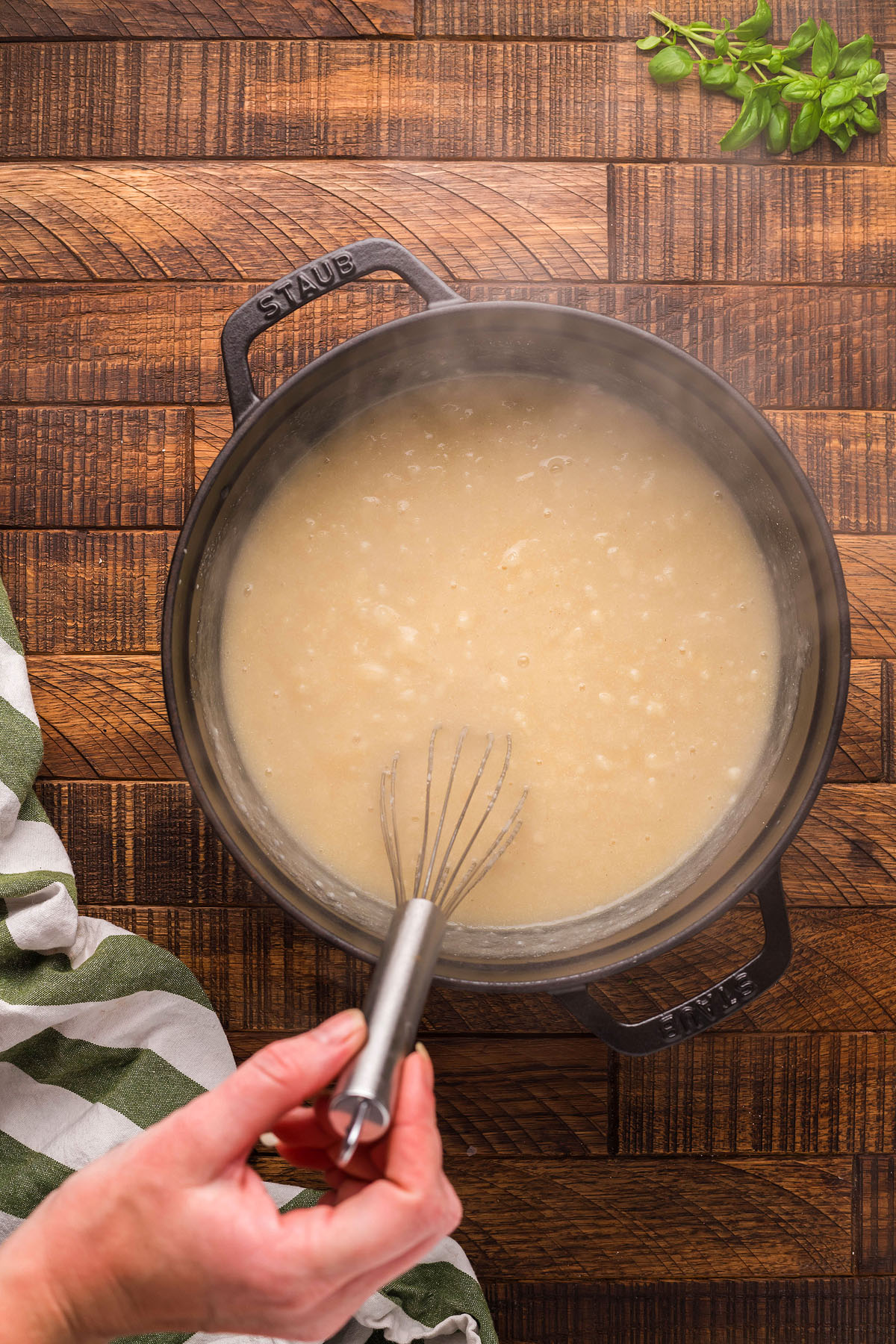 A hand whisks a creamy white sauce in a black pot on a wooden surface, with basil and a striped towel nearby—a perfect start for gluten free meatball recipe or other Italian meatball dinner ideas.