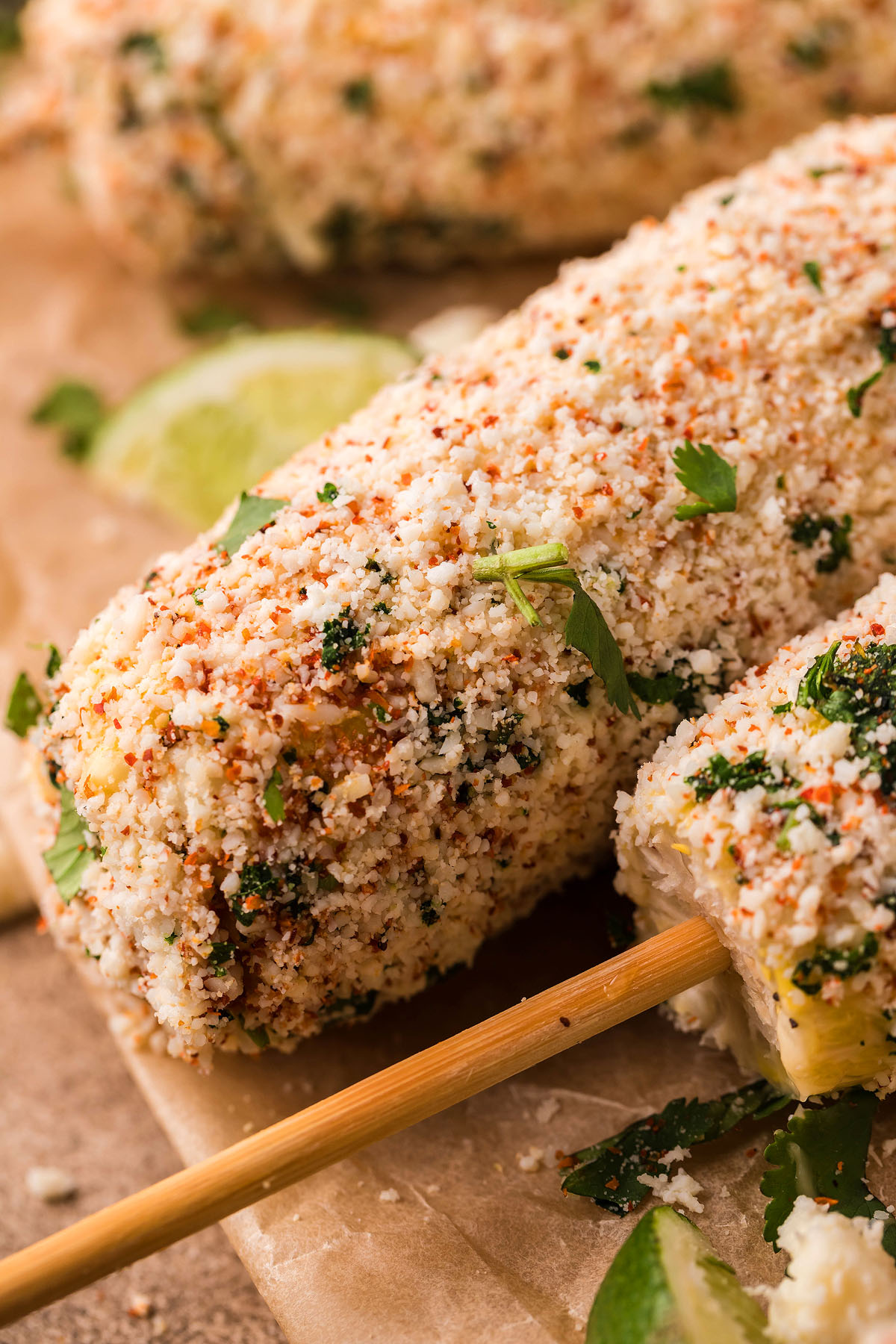 Close-up of Smoked Elote – Mexican Street Corn on a stick, coated with cheese, spices, and herbs, with lime wedges nearby.