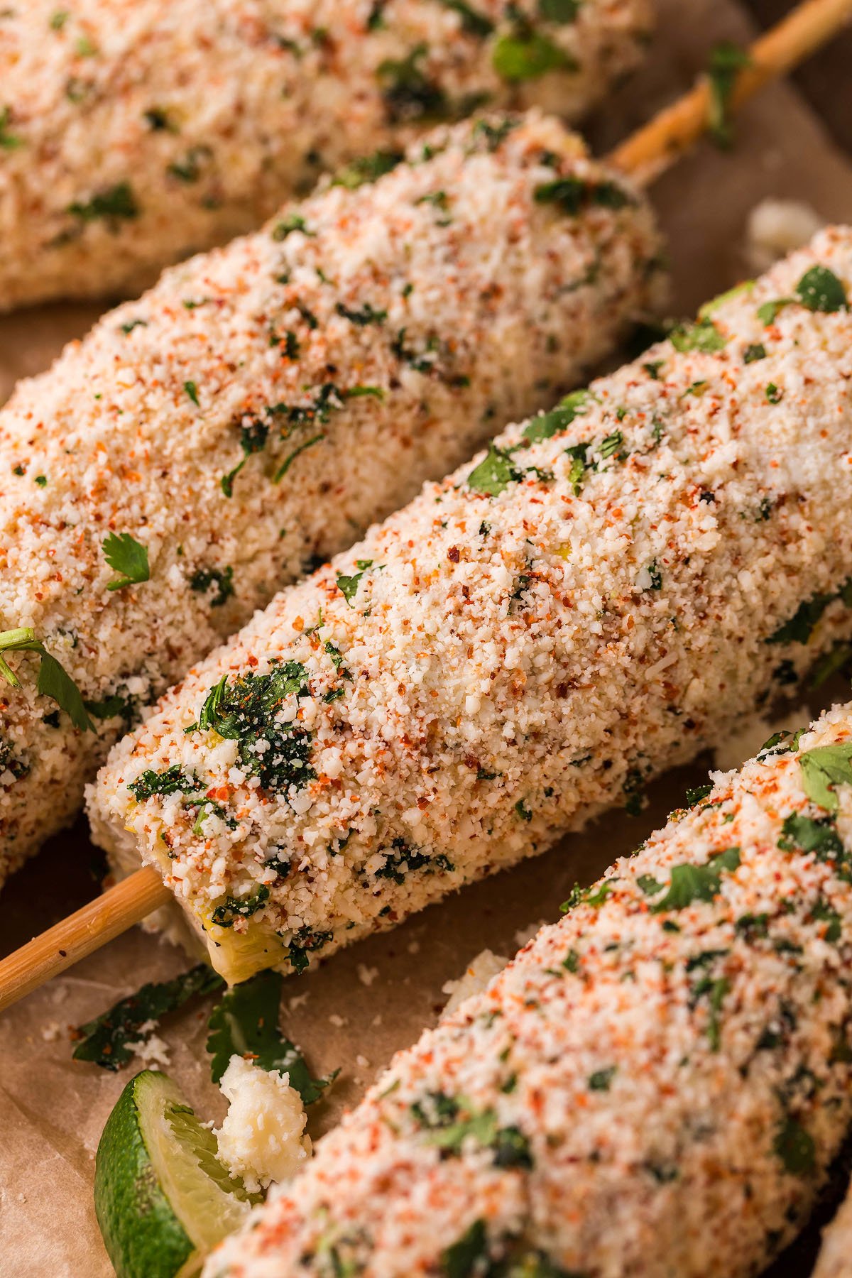 Smoked Elote – Mexican Street Corn on skewers, coated with cheese, spices, and cilantro, served alongside lime wedges for a burst of flavor.