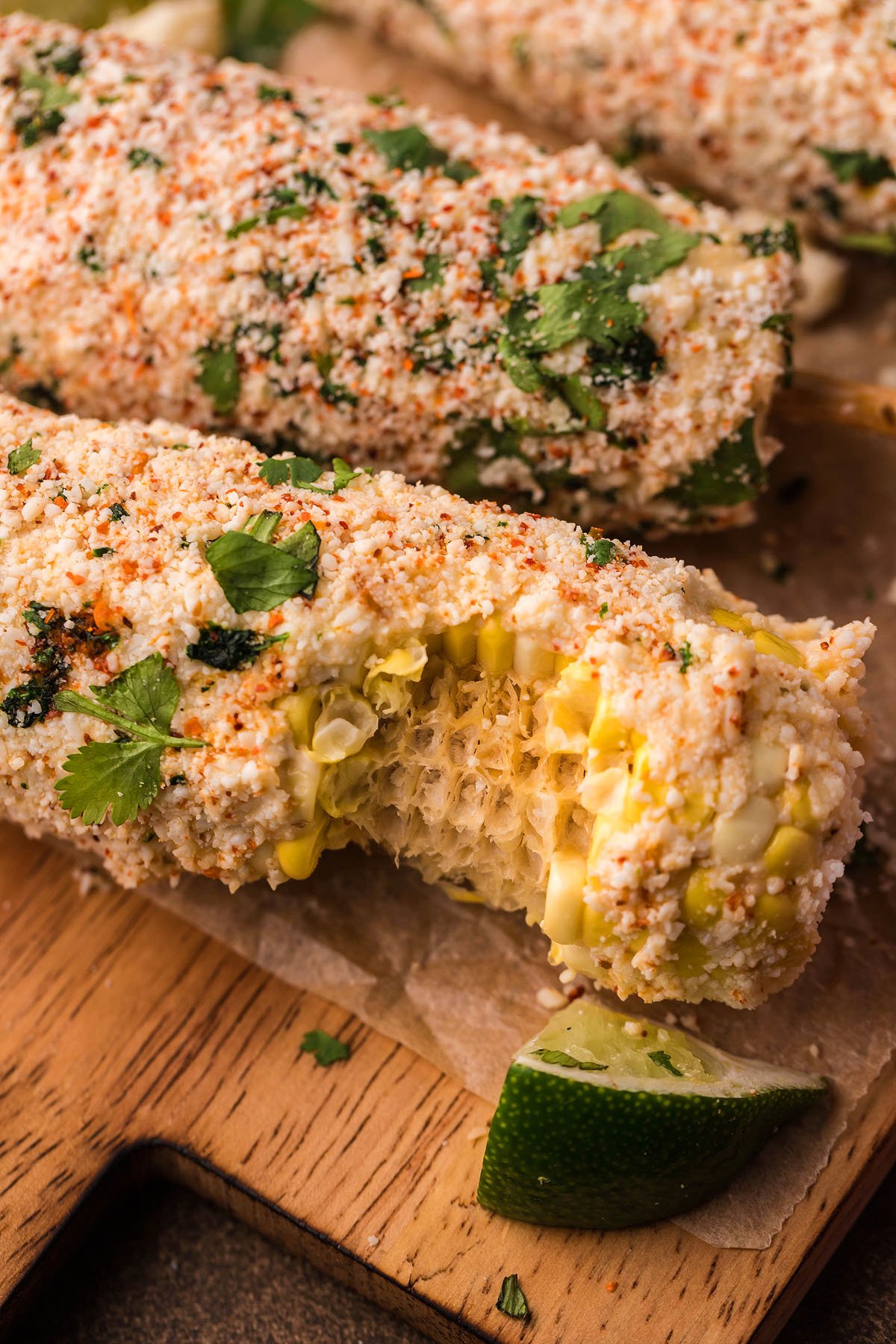 Smoked Elote – Mexican Street Corn on the cob, topped with cheese, spices, and cilantro, served with a lime wedge on a wooden board.