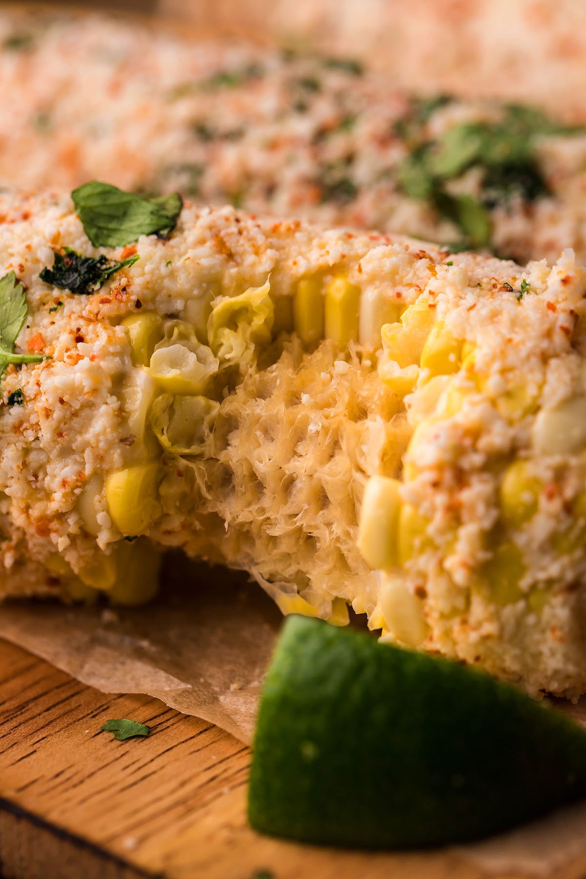 Close-up of Smoked Elote – Mexican Street Corn on the cob, topped with cheese, spices, cilantro, and a lime wedge on a wooden surface.