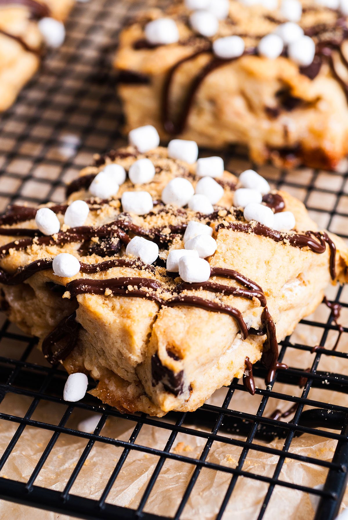A S’more Scones recipe featuring a s'mores-flavored pastry topped with chocolate drizzle, mini marshmallows, and crumbs on a cooling rack.
