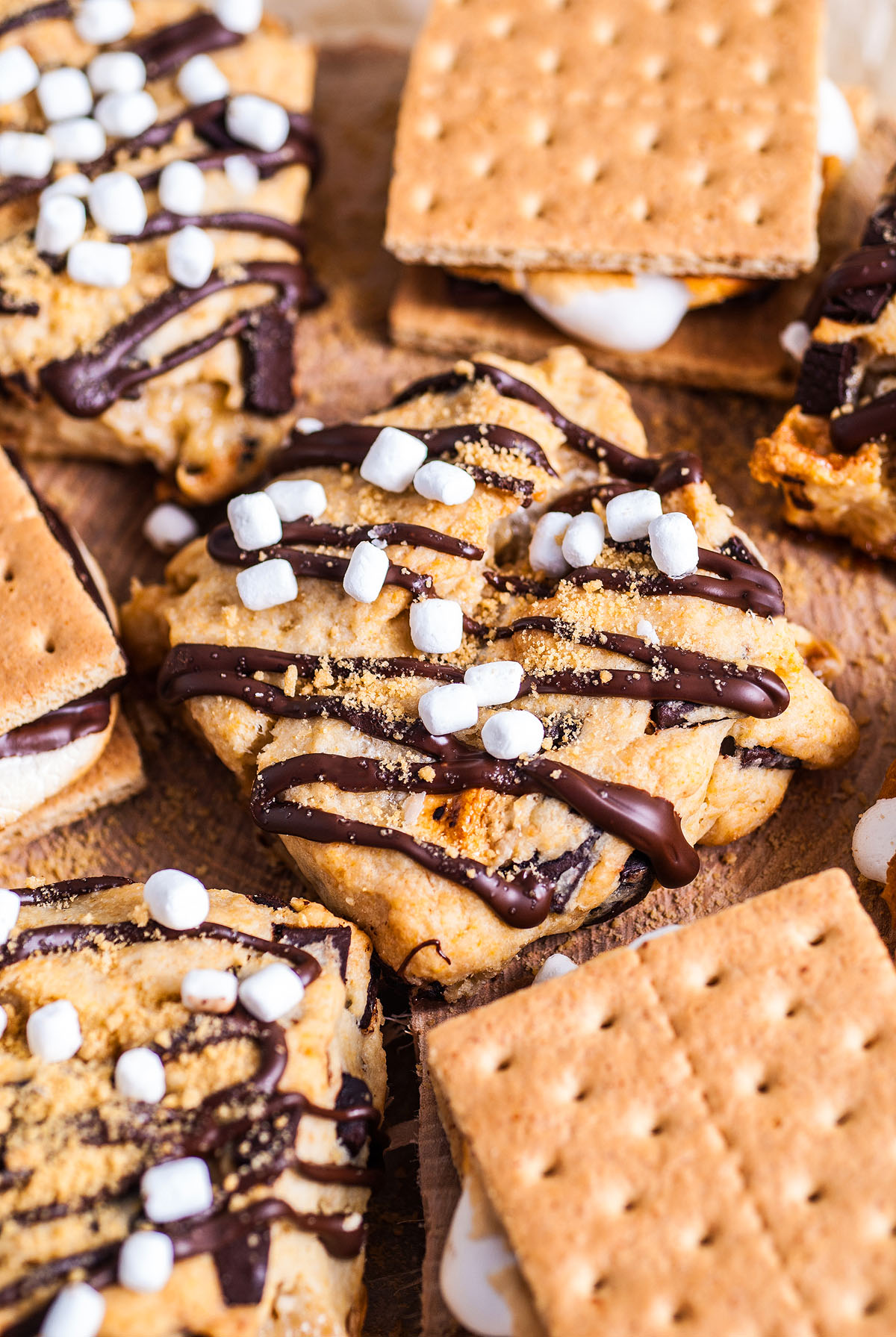 Chocolate-drizzled cookies with mini marshmallows sit beside classic s’mores and irresistible S’more Scones on a wooden surface—a tempting display for any scones recipe lover.
