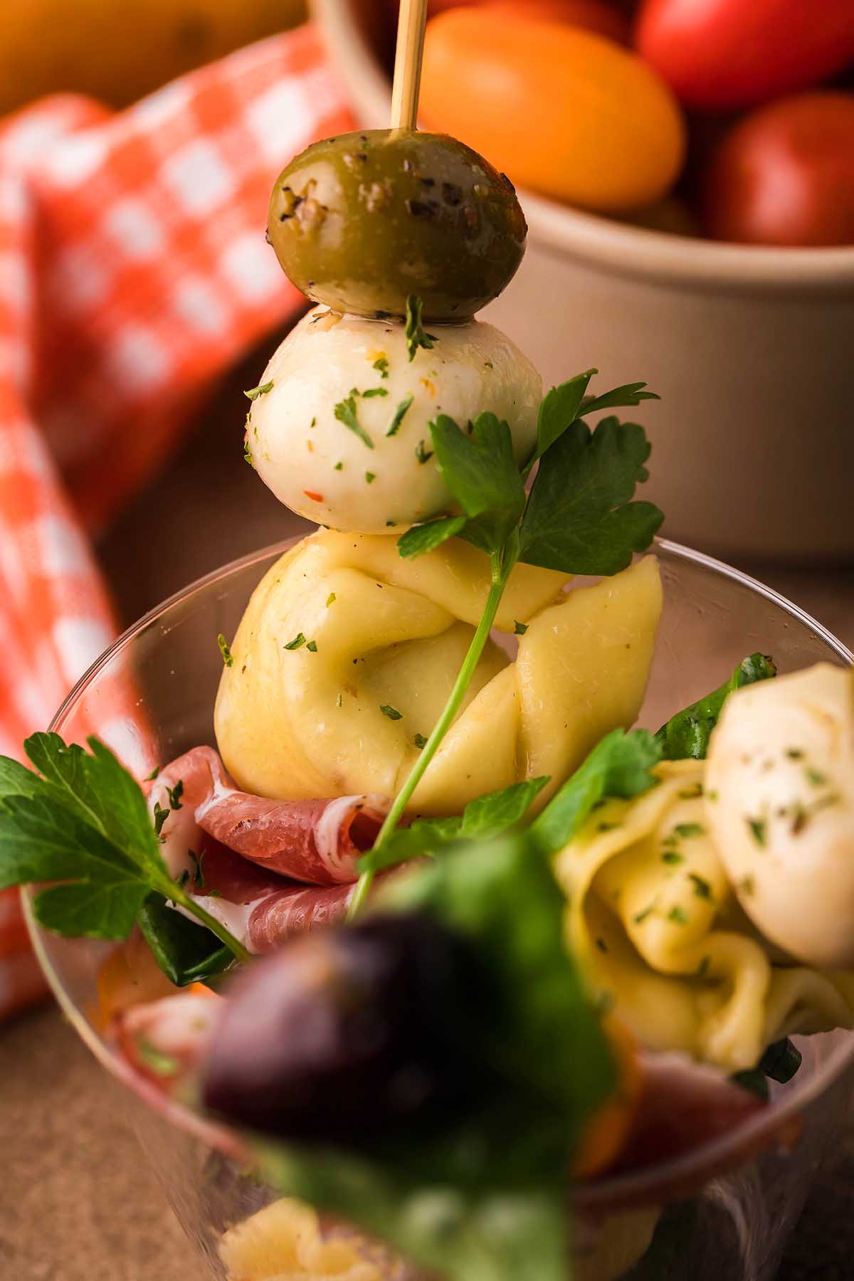 Antipasto skewers with olive, mozzarella, tortellini, and herbs in a clear cup with a checkered napkin in the background&mdash;an easy favorite for Italian appetizer recipes.