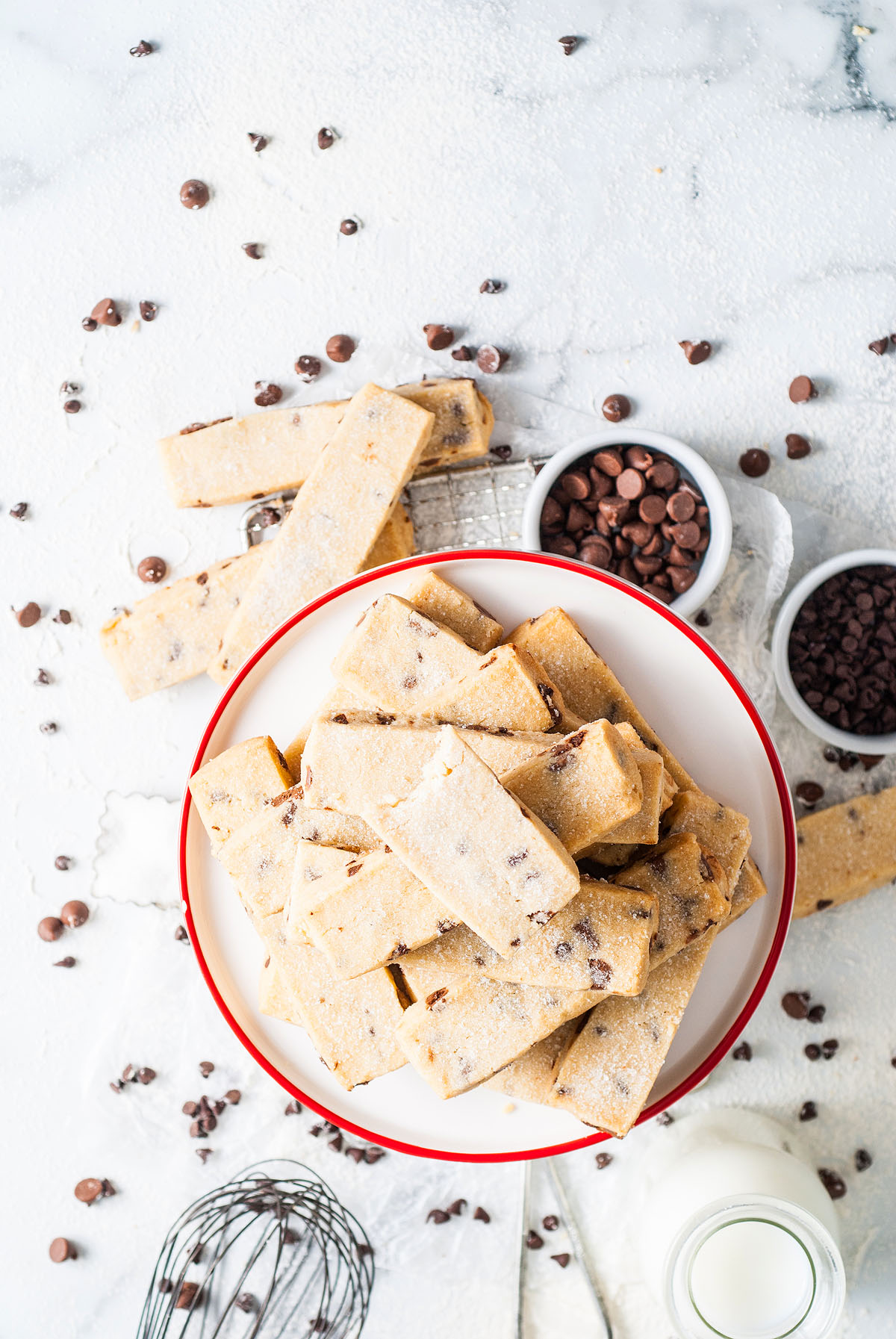 A plate of chocolate chip cookie dough bars, reminiscent of chocolate chip shortbread cookies, is surrounded by chocolate chips and a whisk on a white surface.