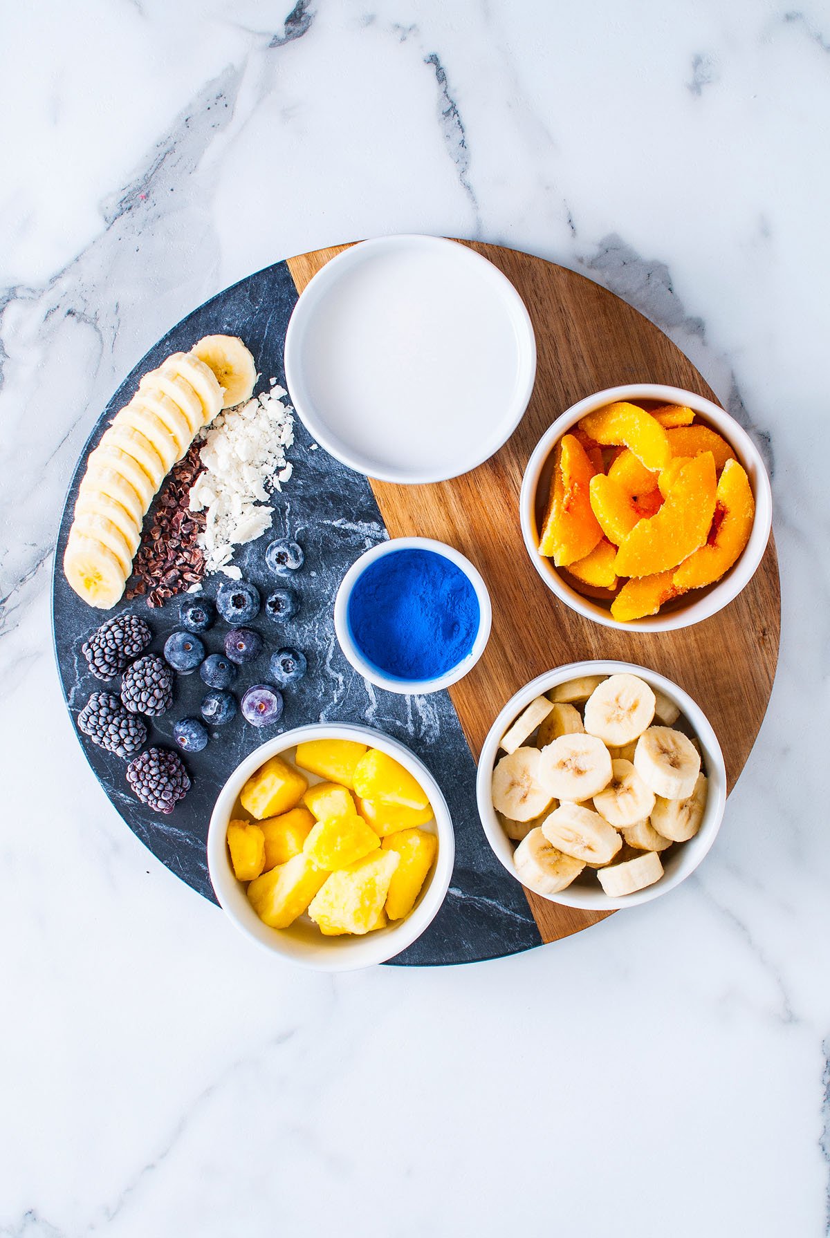 A round board with a Blue Smoothie Bowl with Spirulina, surrounded by bowls of berries, bananas, mango, peach slices, powder, and a small bowl of liquid.