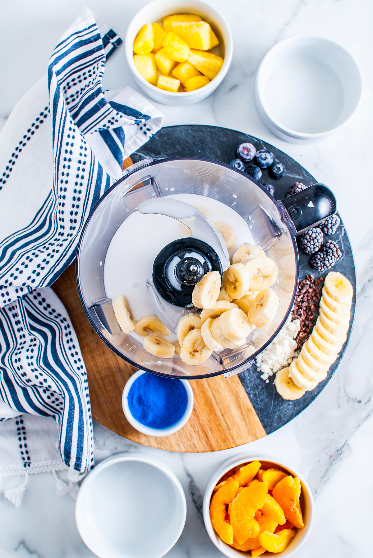 A food processor with bananas and milk, surrounded by fruit bowls—including a Blue Smoothie Bowl with Spirulina—and a striped napkin on a marble surface.