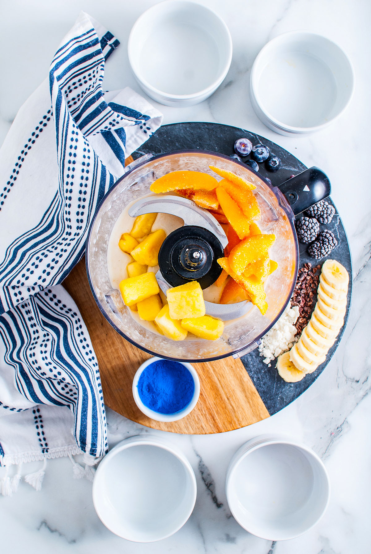 A food processor with mango and pineapple, surrounded by fruit, bowls, a towel, and blue powder ready for a Blue Smoothie Bowl with Spirulina on a marble surface.