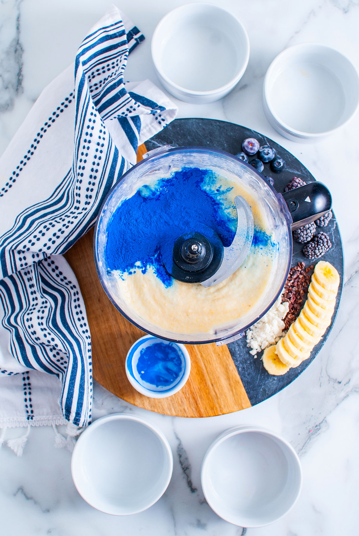 A food processor with blue powder on smoothie mix, perfect for creating a Blue Smoothie Bowl with Spirulina, surrounded by fresh fruit, bowls, and a striped towel.