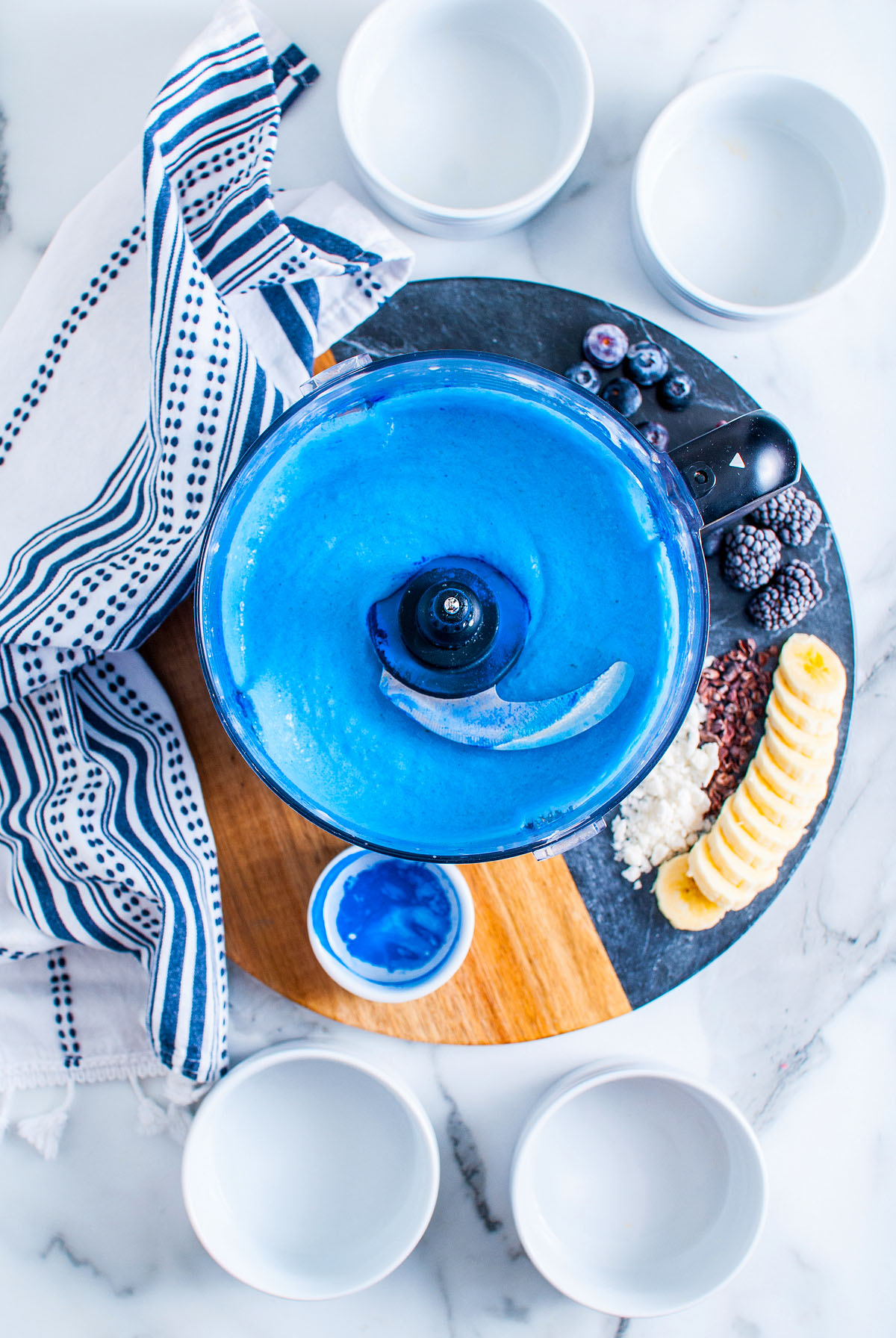 A food processor with a bright blue smoothie, perfect for a Blue Smoothie Bowl with Spirulina, surrounded by bowls, fruit, and a striped cloth on a marble surface.