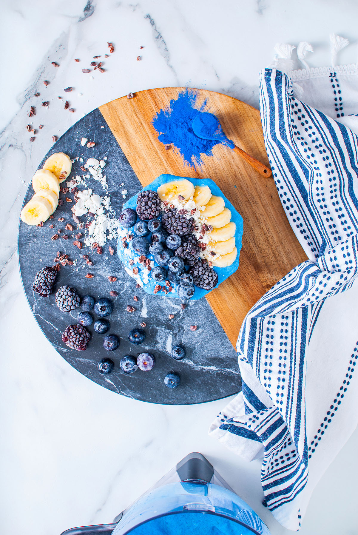 Blue Smoothie Bowl with Spirulina, topped with banana slices, berries, coconut, and blue powder on a round wooden board; striped towel beside.