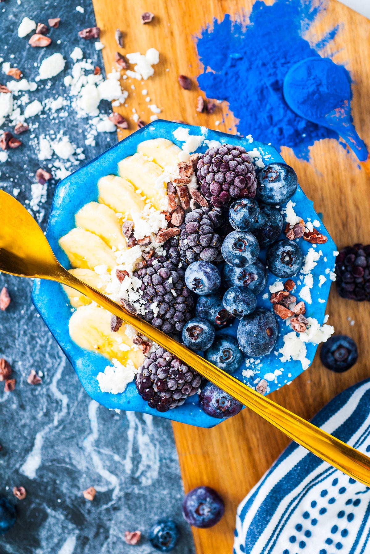 A Blue Smoothie Bowl with Spirulina topped with bananas, blackberries, blueberries, and a gold spoon on a wooden board.