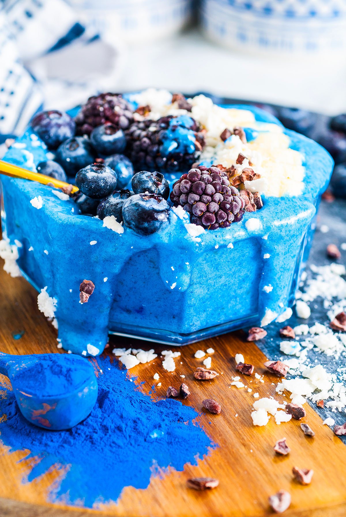 A Blue Smoothie Bowl with Spirulina topped with blueberries, blackberries, coconut, and cacao nibs sits on a wooden board.