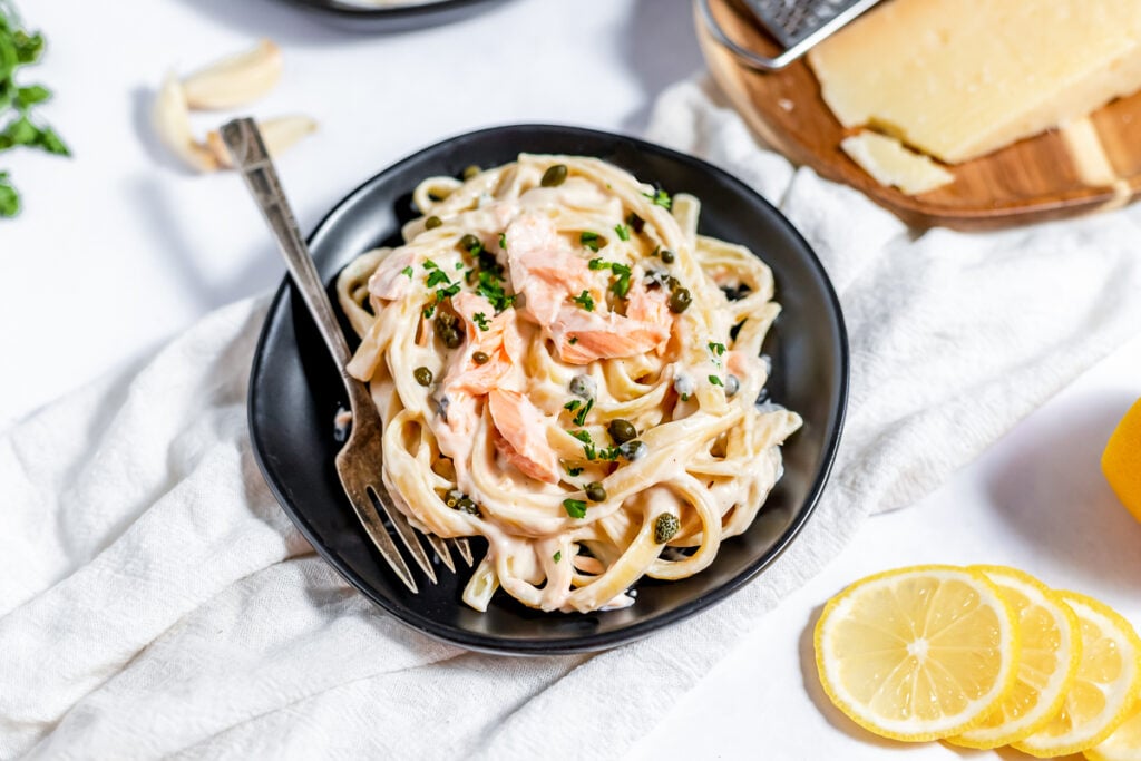 Pasta with creamy sauce, capers and tender salmon chunks on a black plate with a fork
