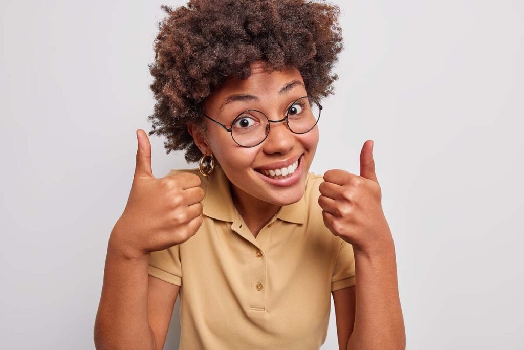 Pleasant looking curly haired woman smiles happily keeps thumbs up.