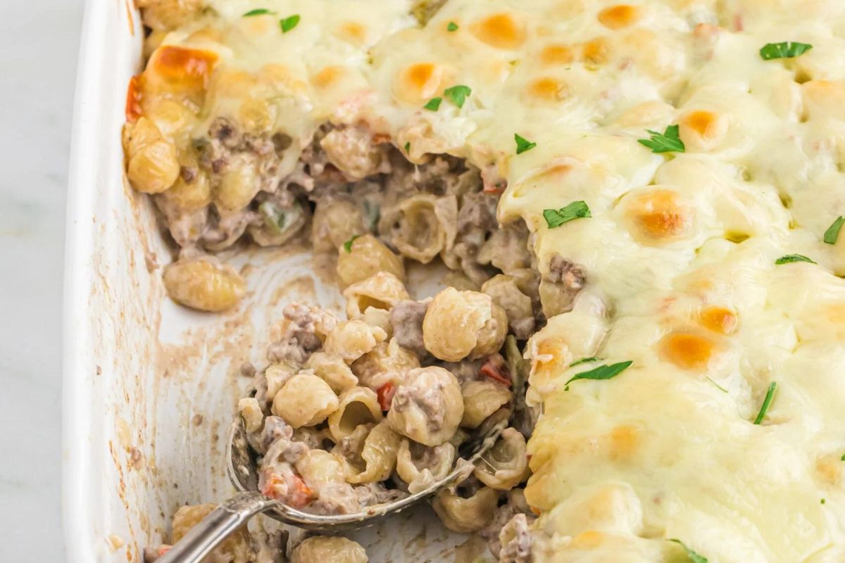 A casserole dish with pasta and thin sliced steak, peppers and onions with plenty of cheese. A serving spoon in the dish.