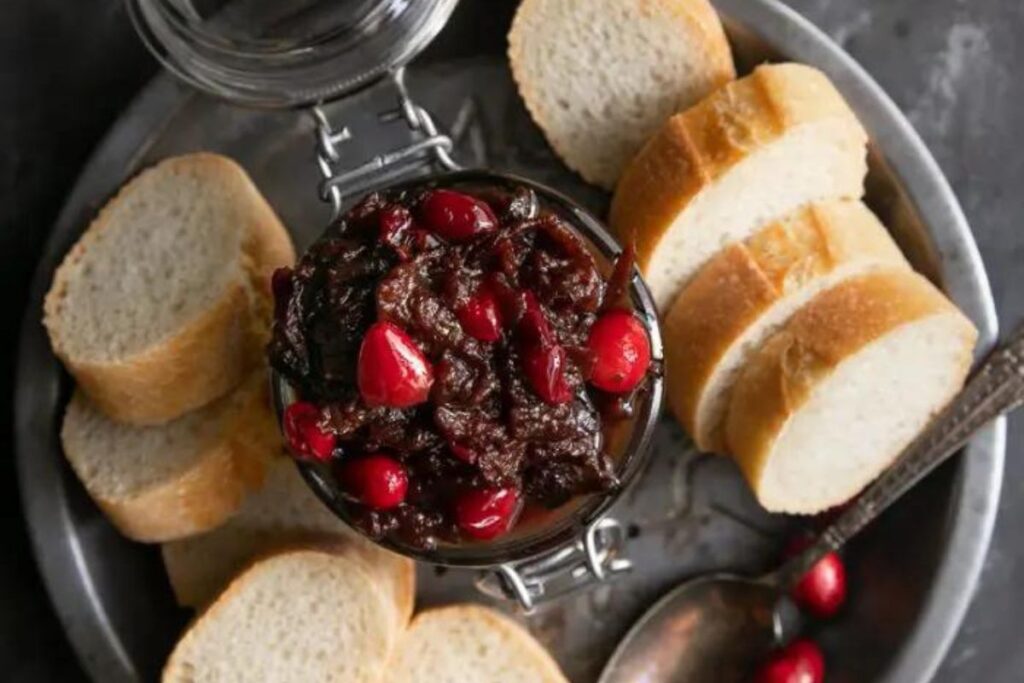 A jar of cranberry bacon jam with bread slices surrounding on a plate.
