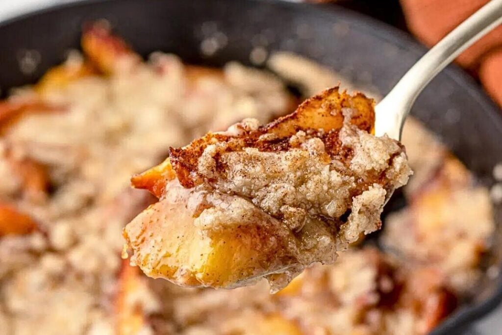 A skillet of peach cobbler with a bite being lifted out.