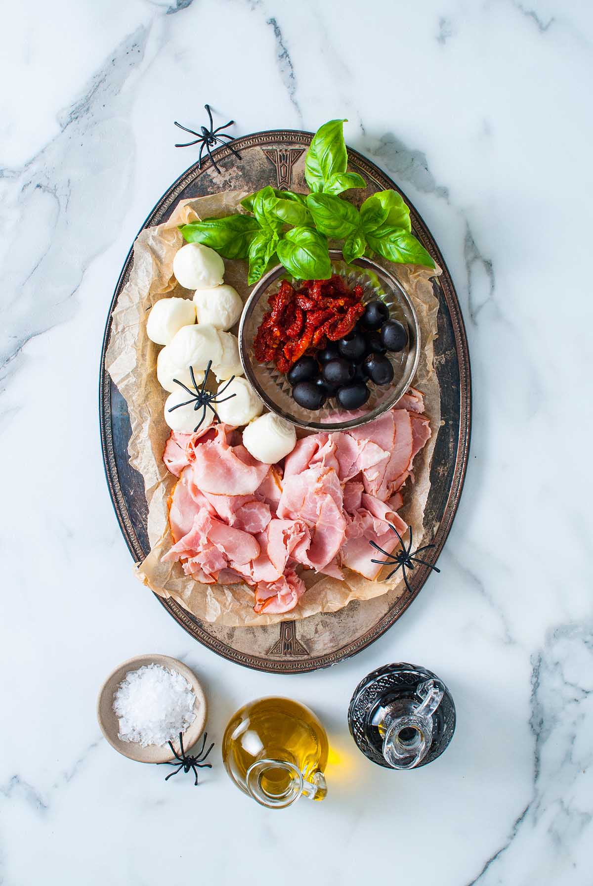 Oval platter with ham, mozzarella, olives, sundried tomatoes, basil, and fake spiders creates a spooky Halloween Antipasto Eyeballs Appetizer on a marble surface.