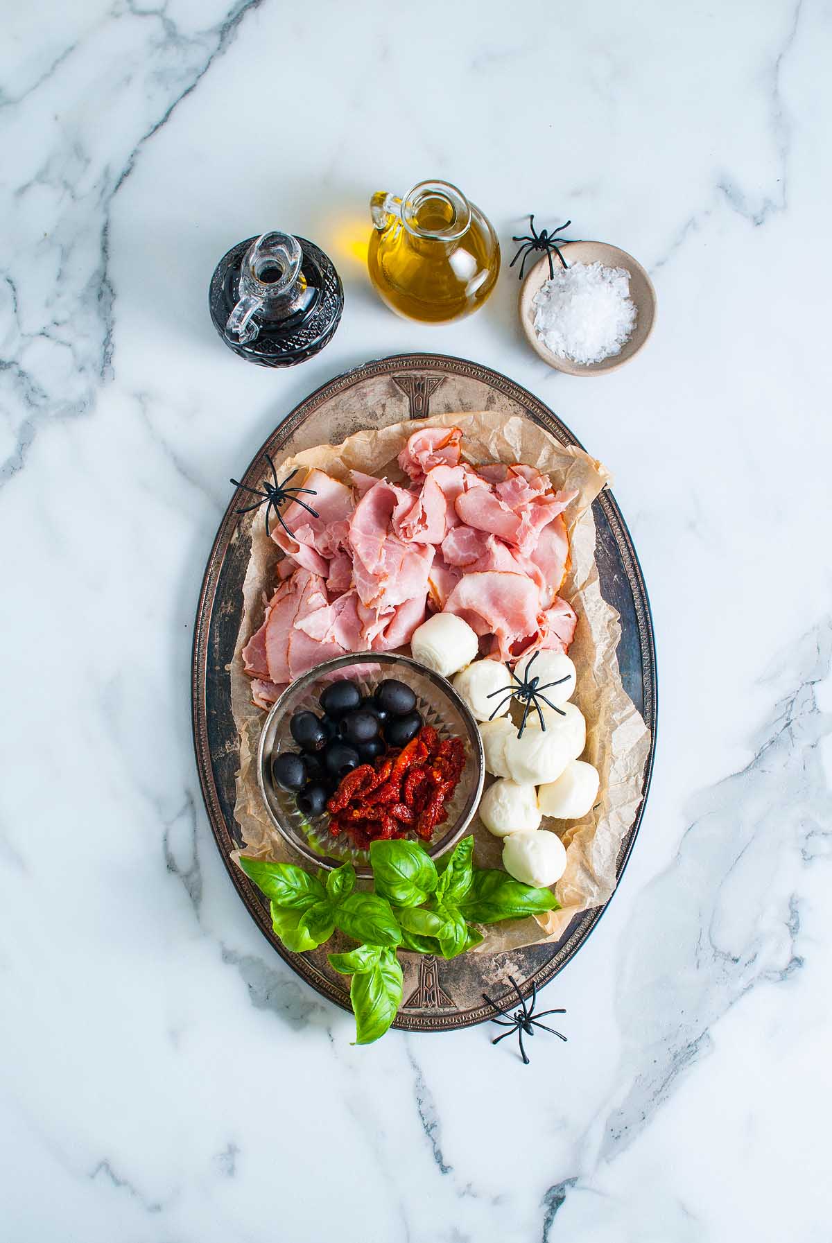 Halloween Antipasto Eyeballs Appetizer: Charcuterie platter with ham, mozzarella, olives, sun-dried tomatoes, basil, and fake spiders on a marble surface for a spooky twist.