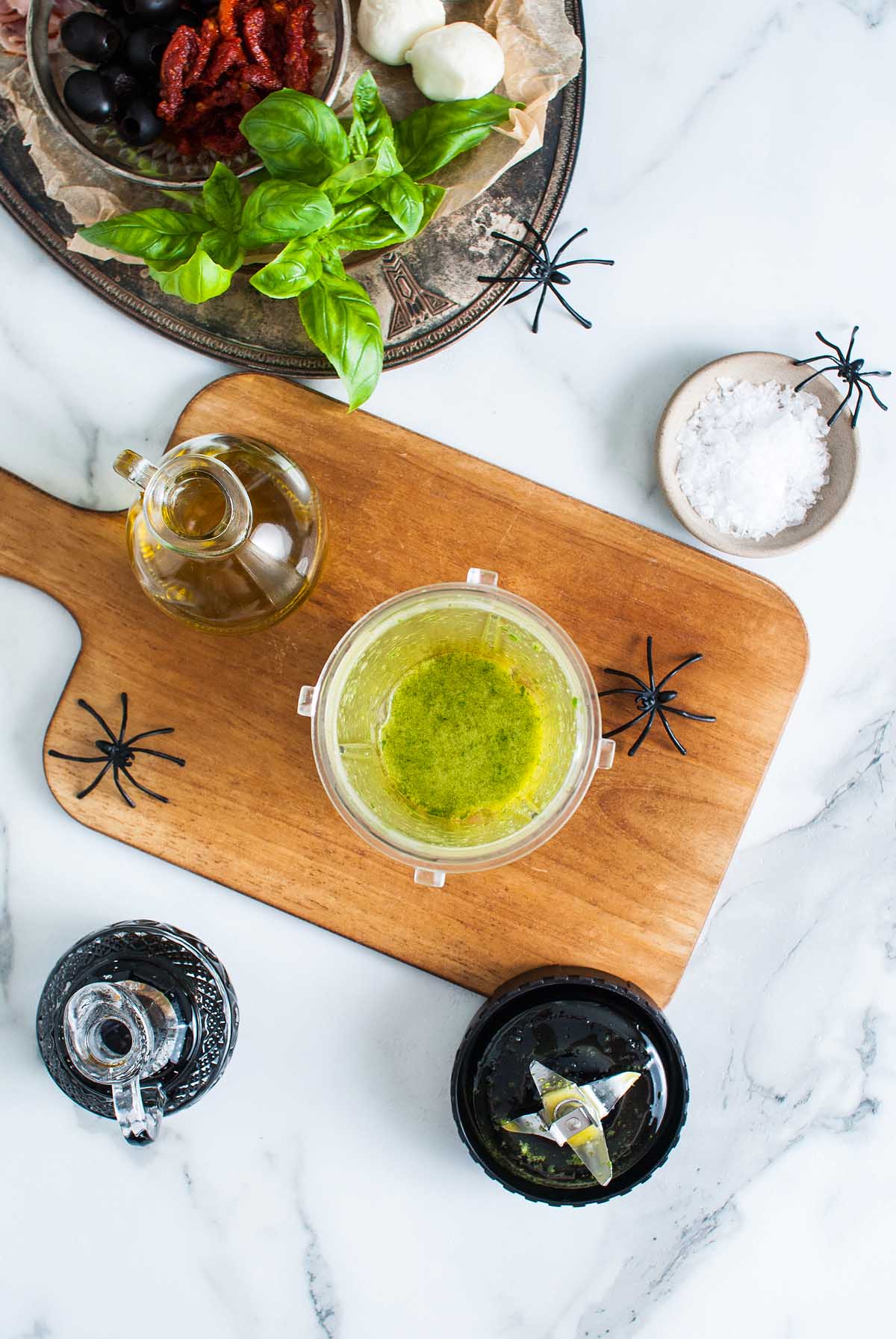 A blender of green sauce on a wooden board with fake spiders, oil, salt, herbs, and sundried tomatoes nearby—perfect for serving alongside your spooky Halloween Antipasto Eyeballs Appetizer.