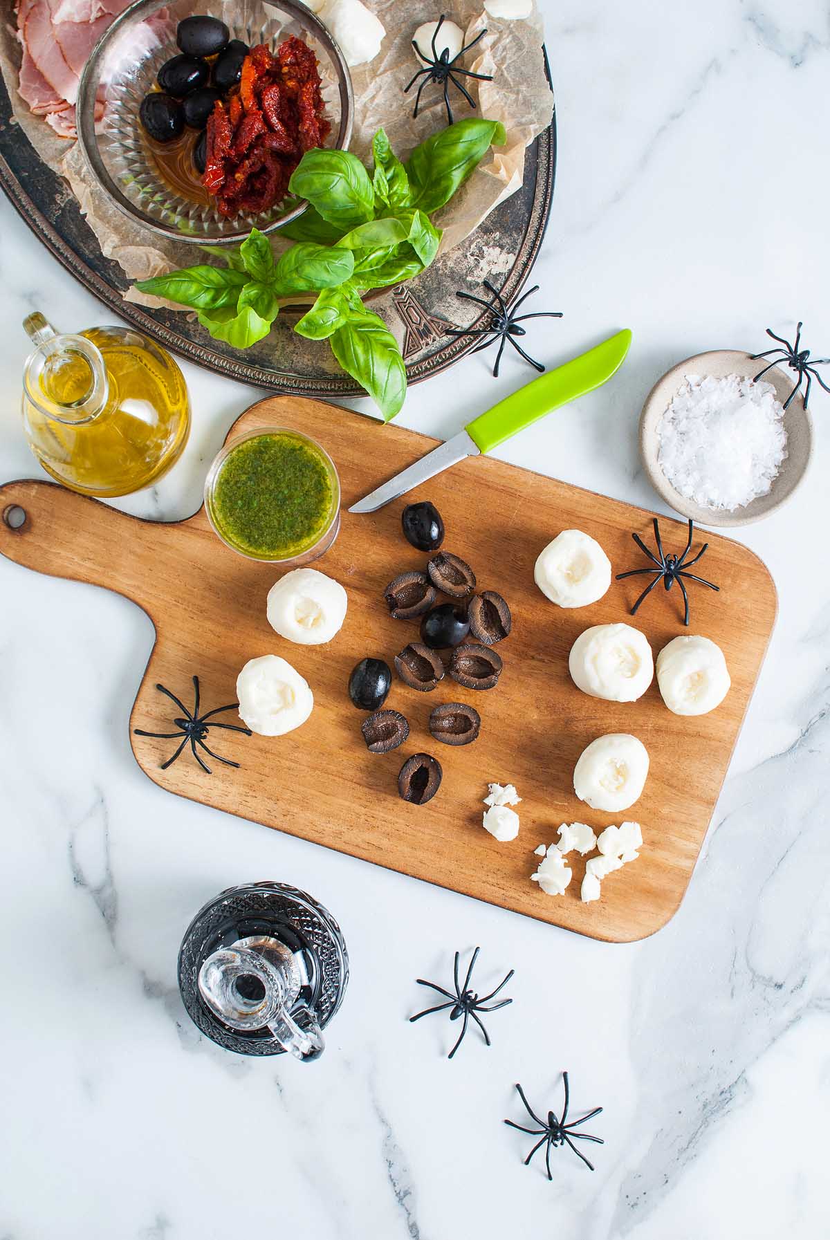 Mozzarella balls and sliced olives on a cutting board create a spooky Halloween Antipasto Eyeballs Appetizer, with fake spiders, pesto, basil, and other ingredients nearby.