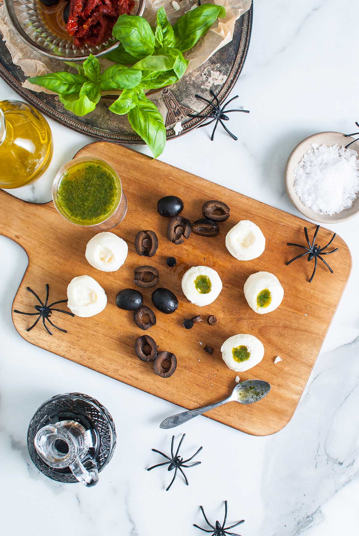 Halloween Antipasto Eyeballs Appetizer: Mozzarella balls with pesto, black olives, and fake spiders on a wooden board, served with herbs and olive oil nearby.