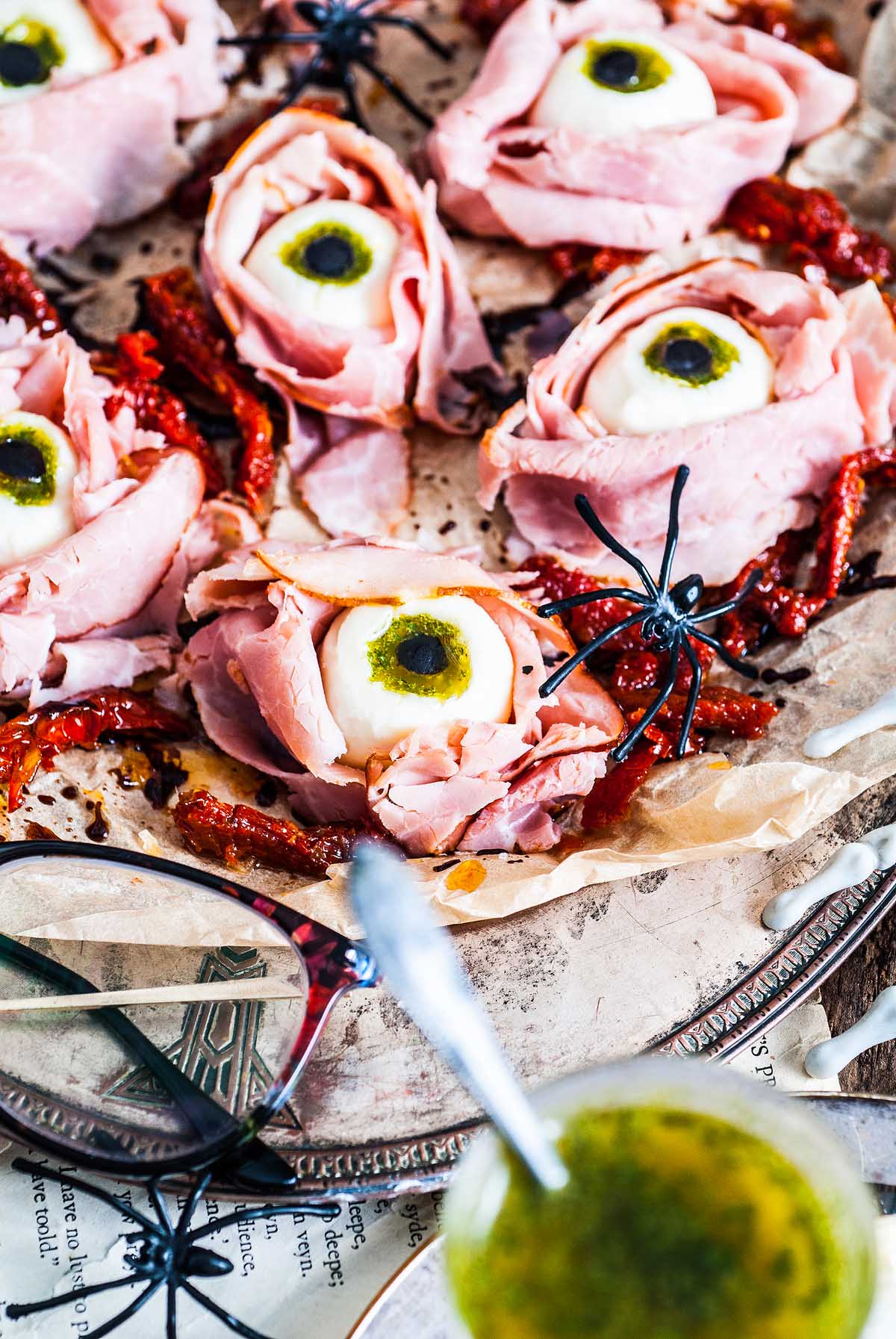Slices of ham arranged as eyes with mozzarella and olives, surrounded by fake spiders and sundried tomatoes, make this the perfect Halloween Antipasto Eyeballs Appetizer.
