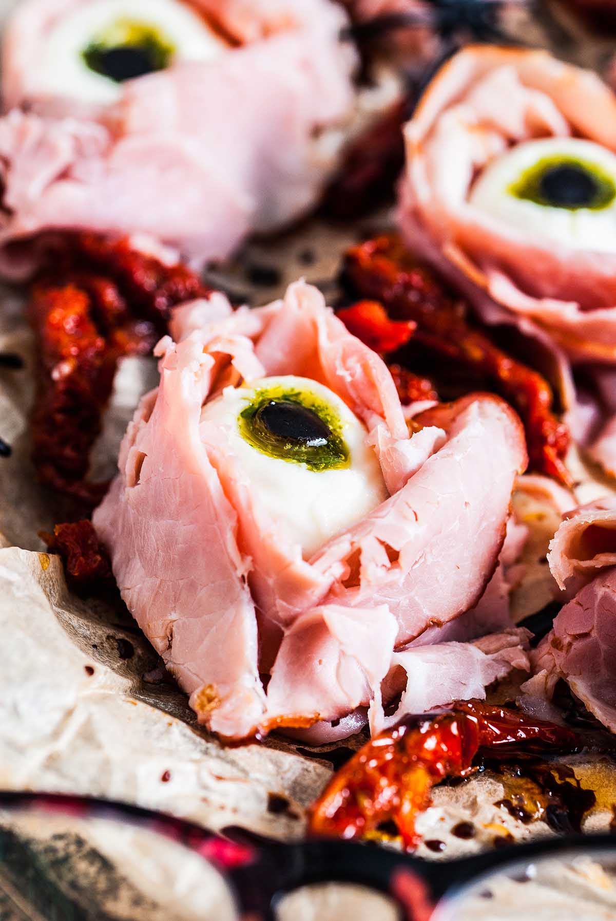Slices of ham arranged to look like Halloween Antipasto Eyeballs, with cheese and an olive in the center, on a tray with sundried tomatoes—a spooky appetizer for your festive spread.