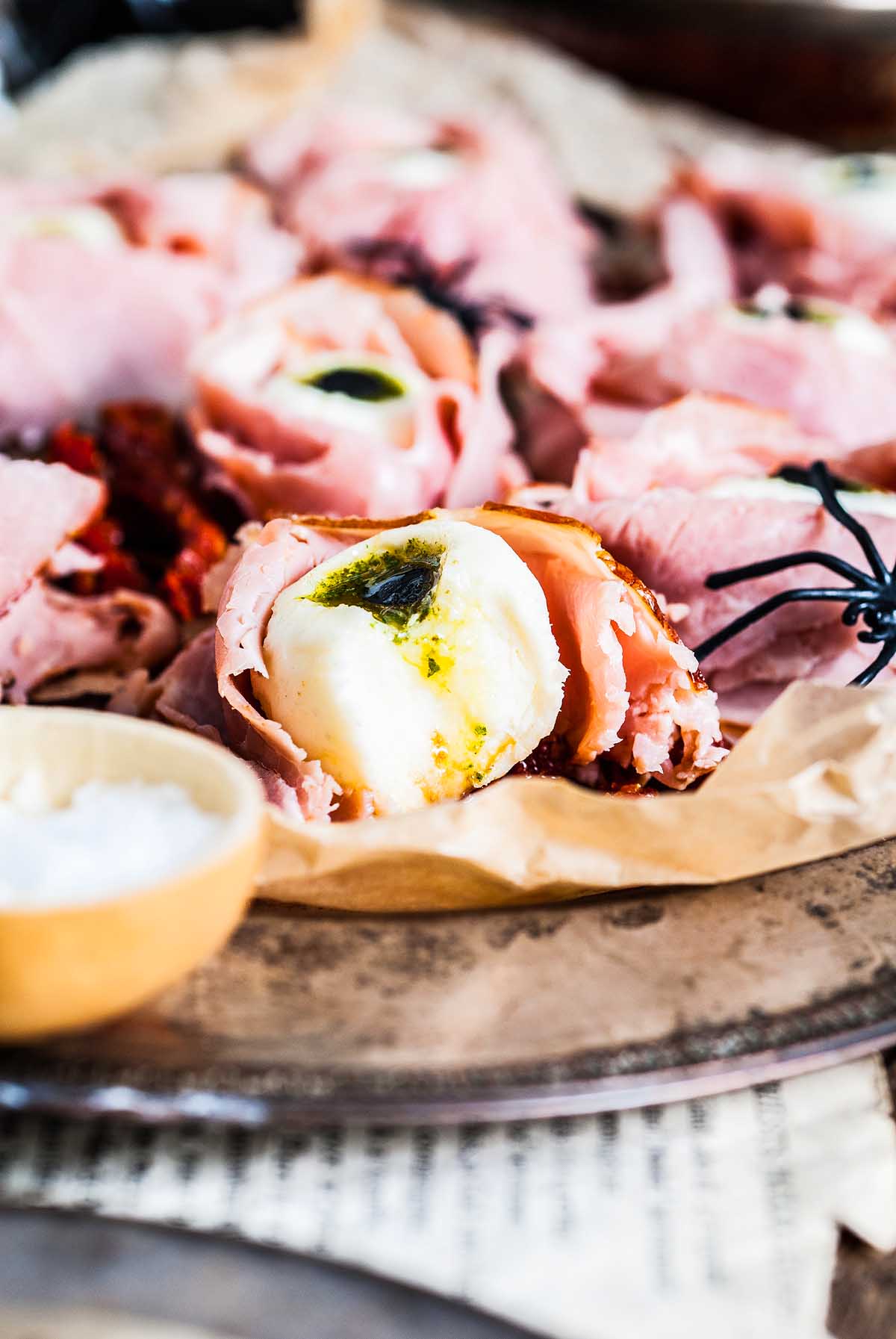 Halloween Antipasto Eyeballs Appetizer: Slices of ham wrapped around fresh mozzarella balls with pesto, served on a rustic platter for a spooky, delicious treat.