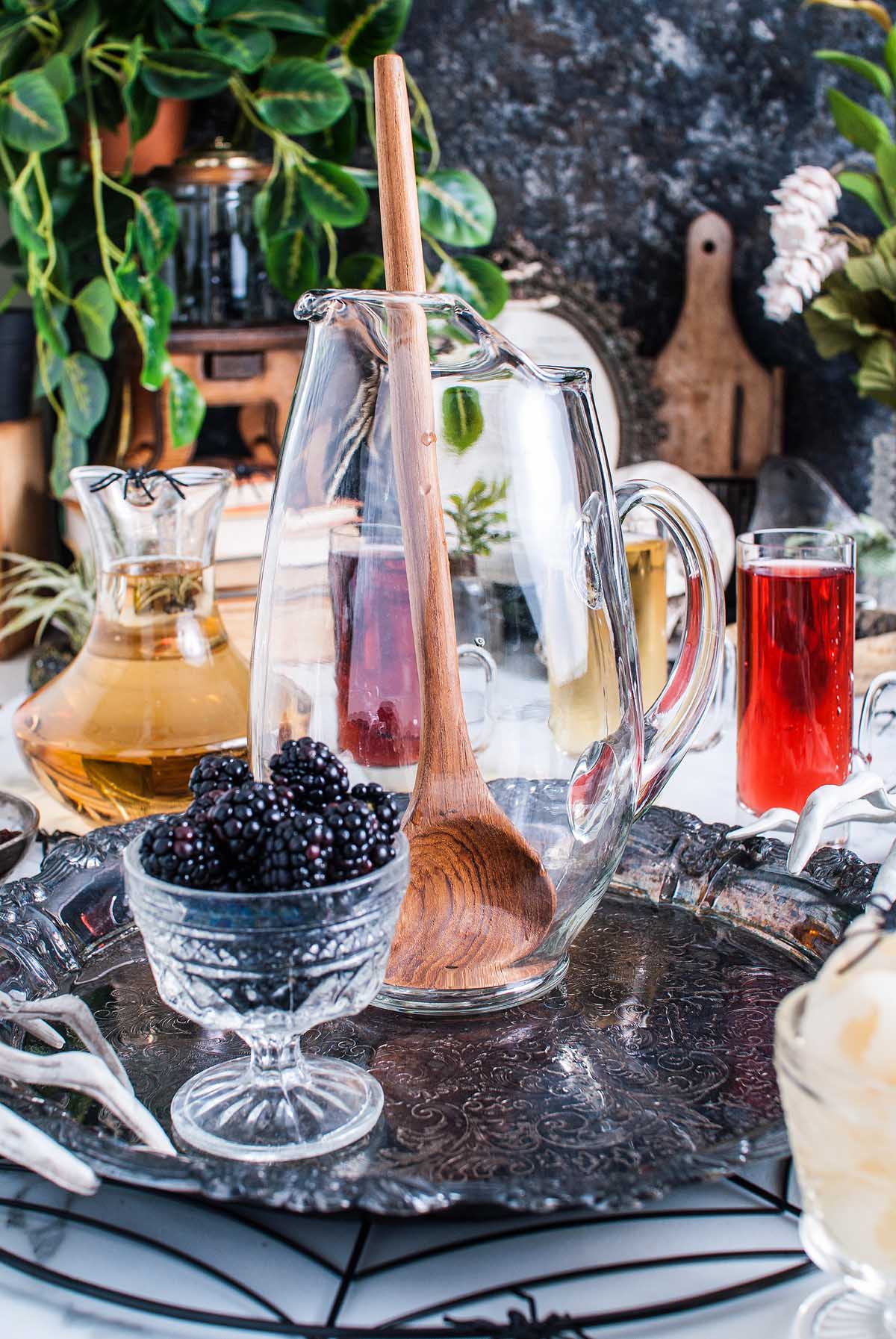 A glass pitcher with a wooden spoon, a bowl of blackberries, and Halloween eyeball punch served on a decorative tray.