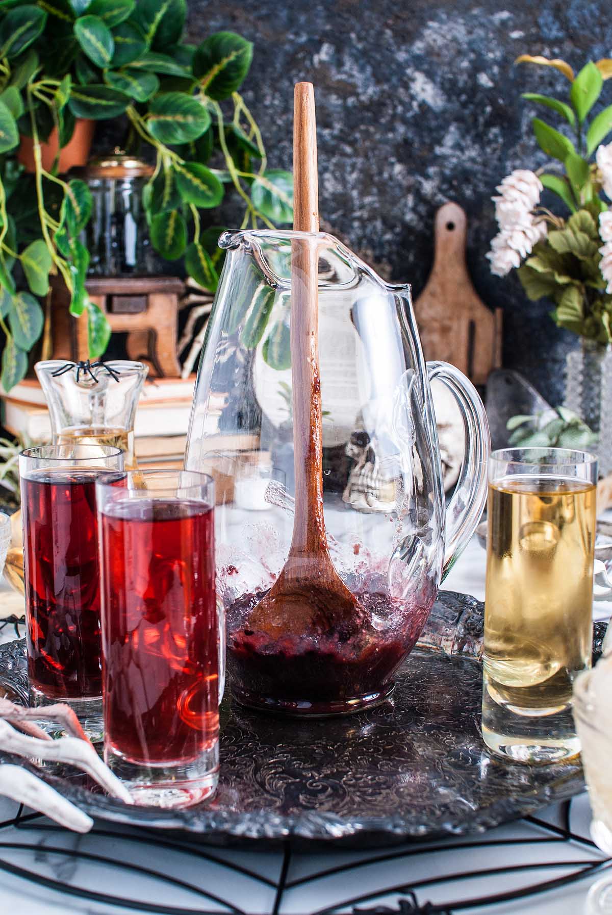 A glass pitcher with a wooden spoon and red drink, surrounded by glasses of red and yellow liquids—perfect for serving your Halloween eyeball punch.