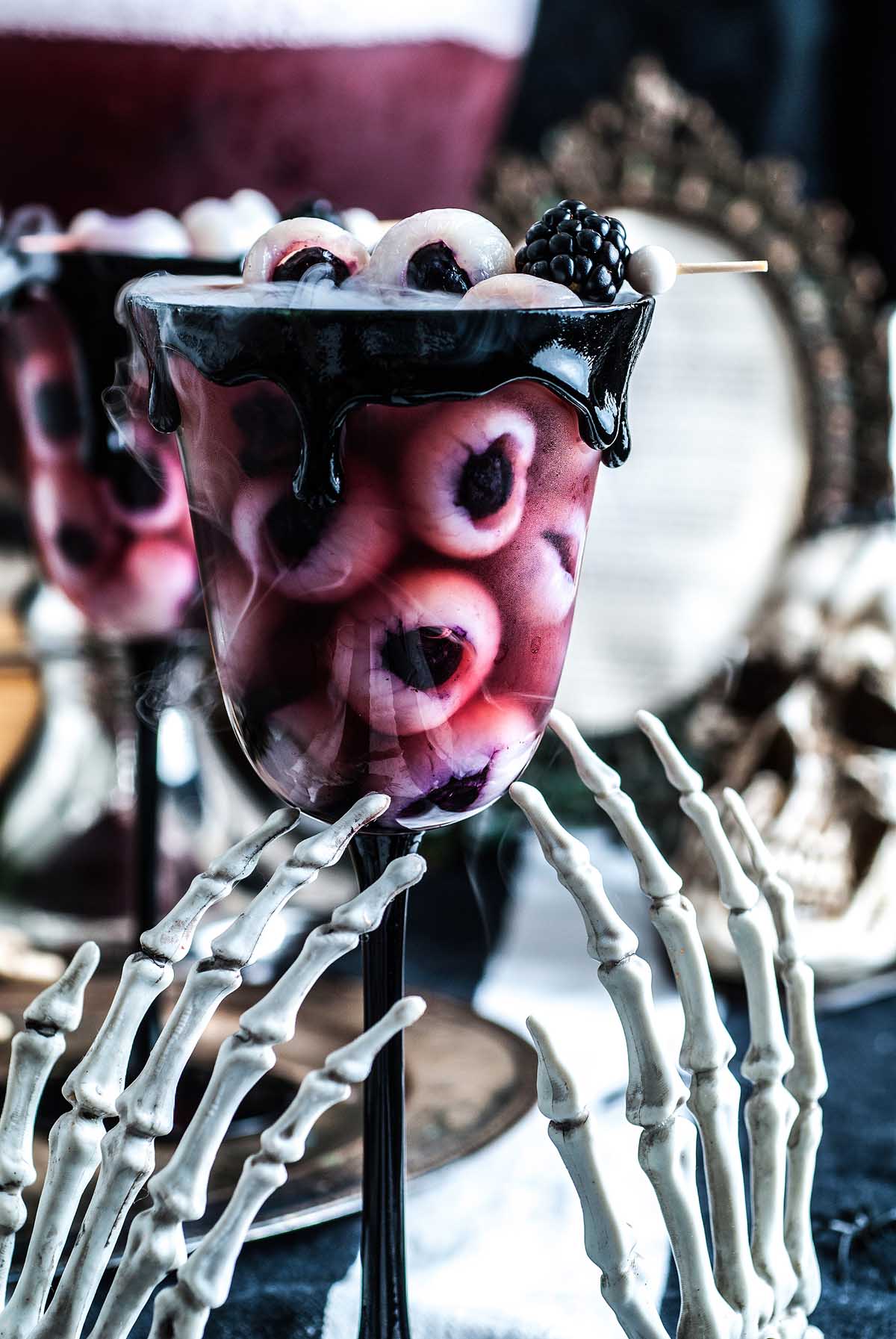A spooky Halloween eyeball punch served in a cocktail glass with eyeball-like fruits and black syrup, eerily held by a skeleton hand.