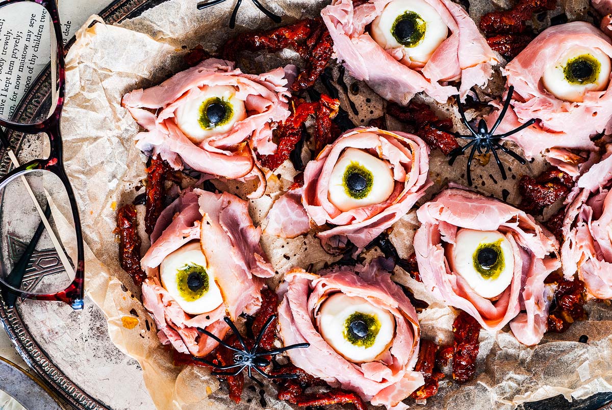 A tray with a spooky Halloween-themed dish featuring ham slices arranged to look like creepy eyes. The center of each "eye" has a round white object resembling an eyeball with a black pupil, surrounded by dry tomato pieces. Fake black spiders are placed as decorations.