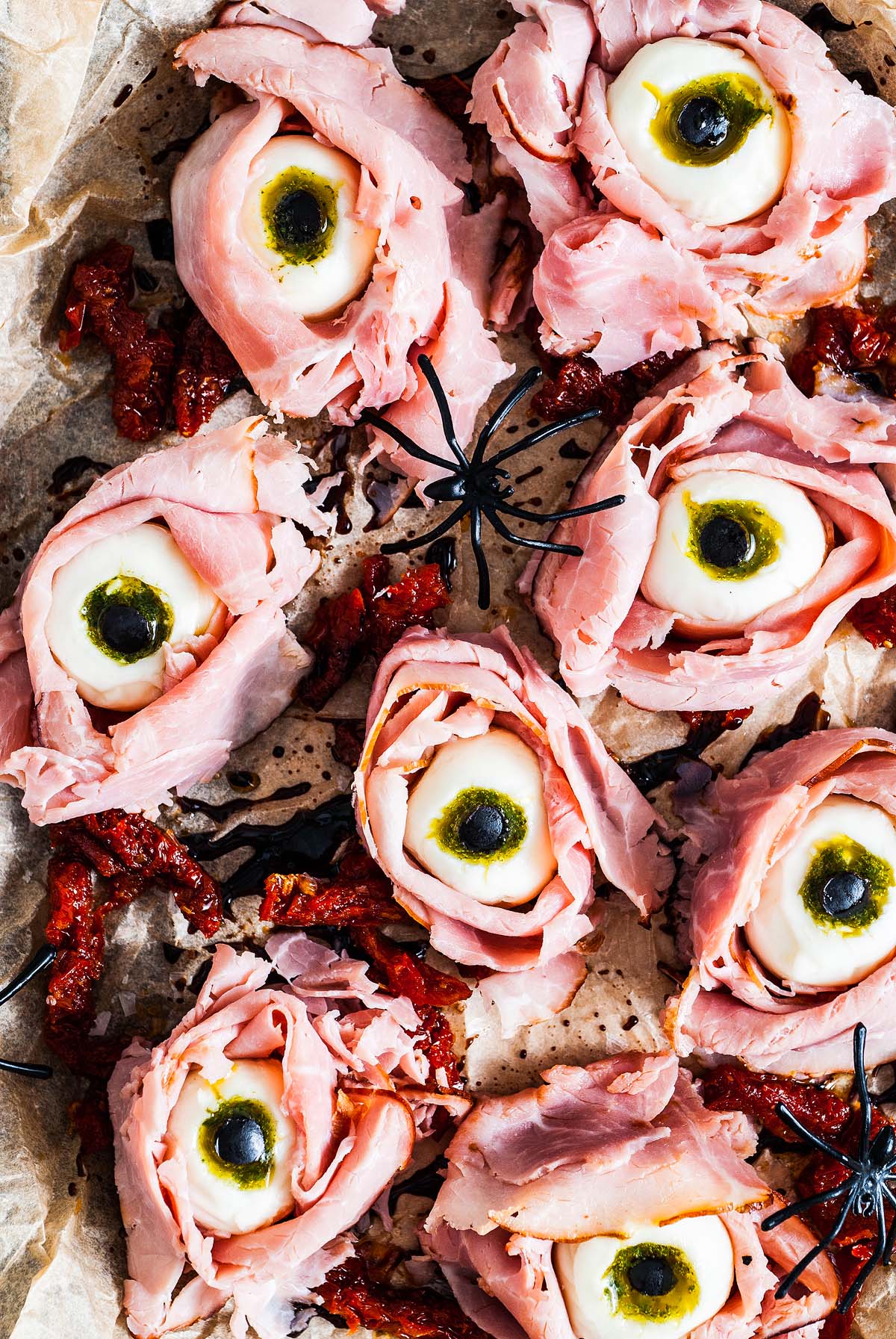 Slices of ham shaped like eyes with mozzarella and olives, arranged on parchment with plastic spiders—these Halloween Antipasto Eyeballs Appetizer are a spooktacular treat for your Halloween party table.