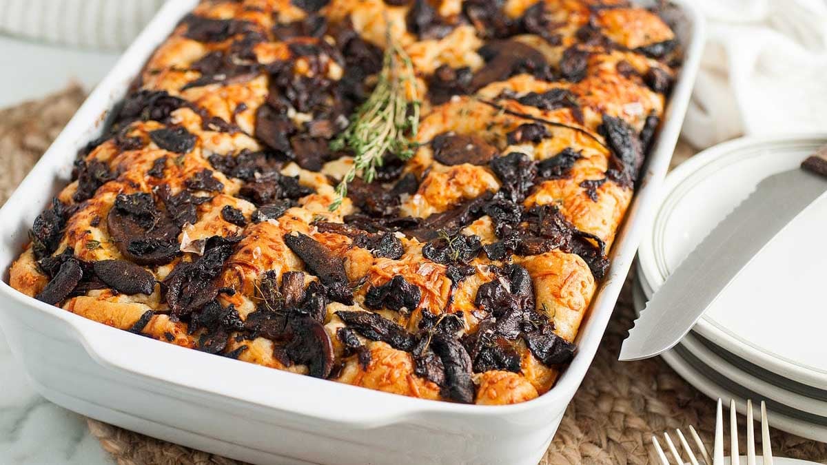 Baked bread pudding topped with mushrooms and herbs in a white casserole dish, perfect for those seeking delicious mushroom recipes, served beside plates and cutlery.