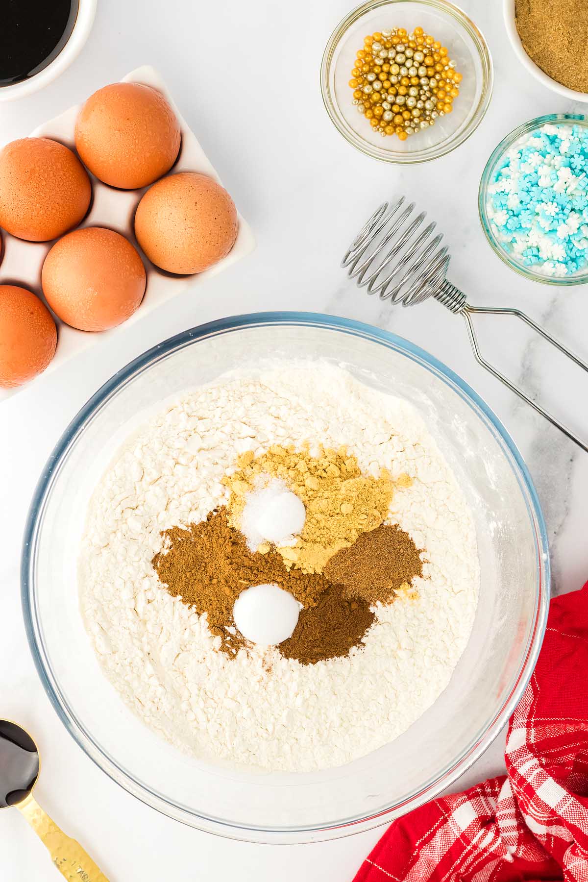 A bowl of flour with spices, eggs, sprinkles, a whisk, and a red towel on a white surface—perfect ingredients for baking 3D Gingerbread Tree Cookies.