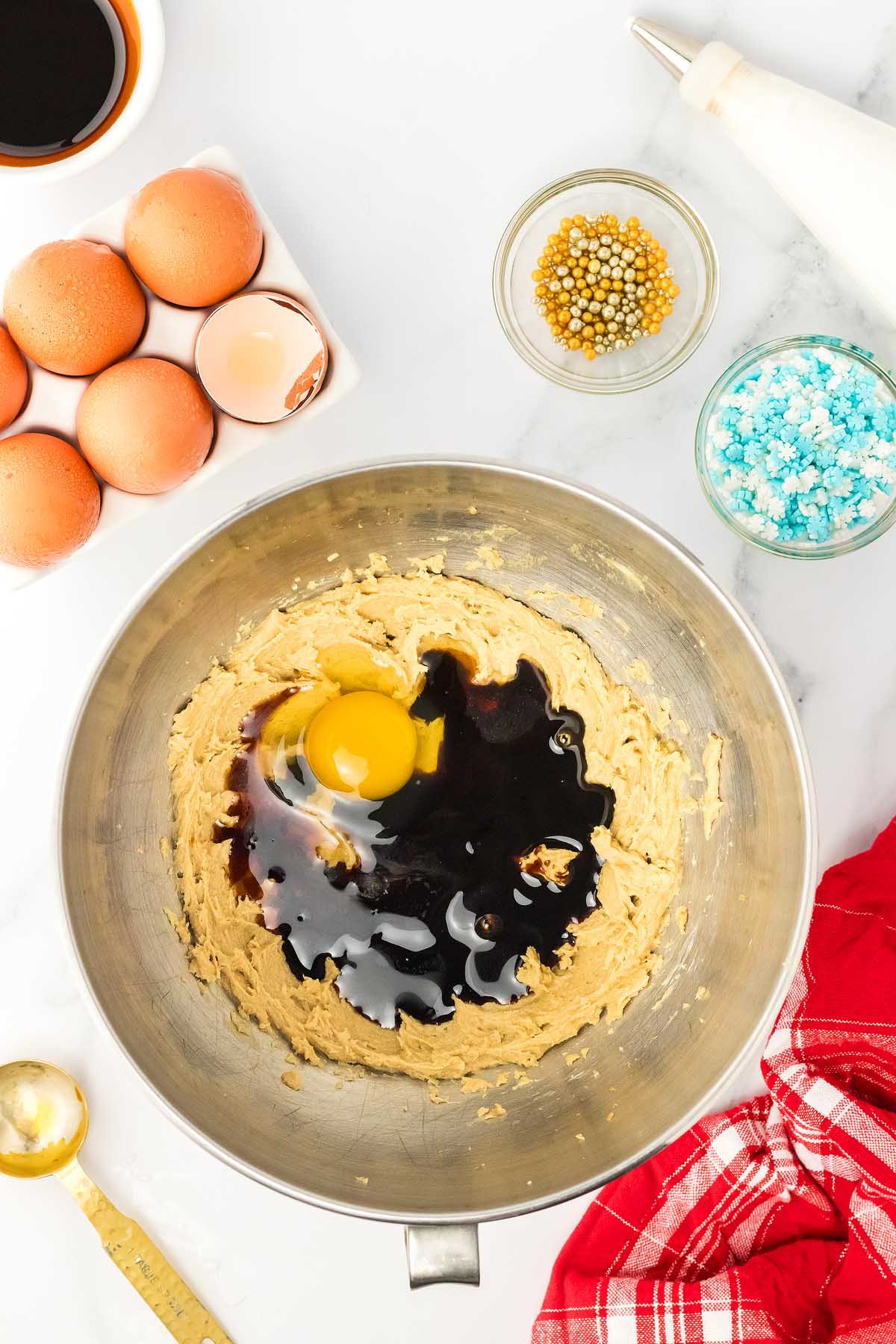 A mixing bowl with cookie dough, an egg, and vanilla sits ready to create 3D Gingerbread Tree Cookies; eggs, sprinkles, and a piping bag are nearby on the counter.