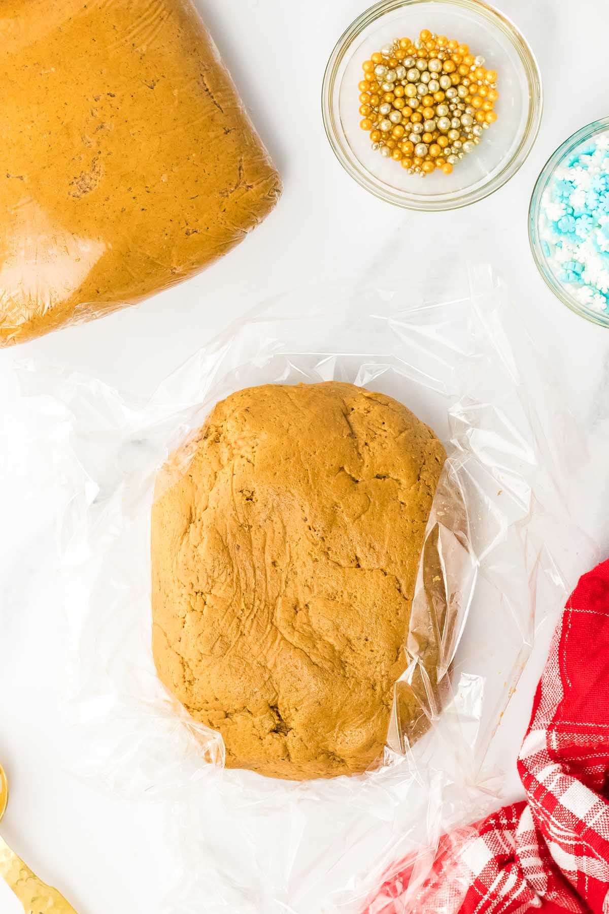 Gingerbread dough on plastic wrap, with sprinkles and another dough piece nearby, ready to be shaped into festive 3D Gingerbread Tree Cookies on a white surface.