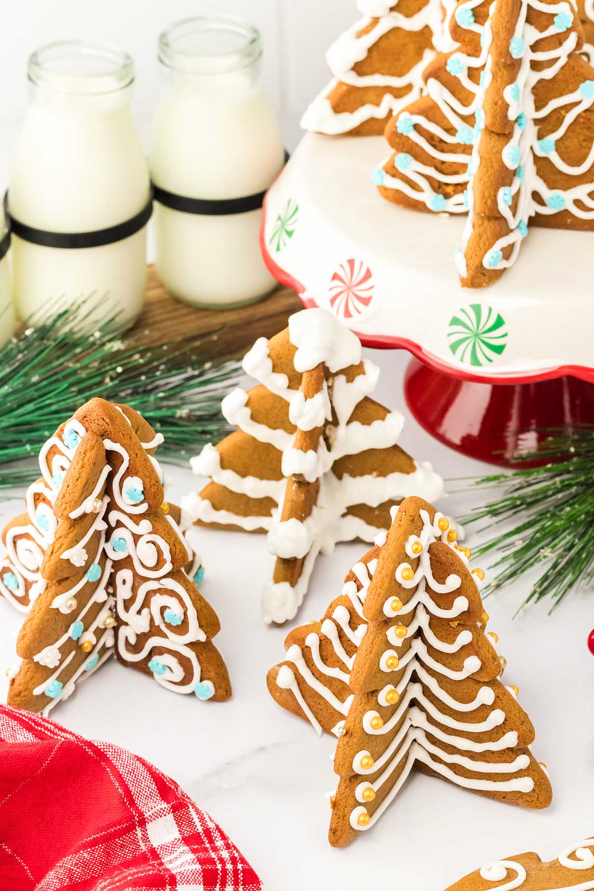 3D Gingerbread Tree Cookies decorated with icing sit on a white table, surrounded by milk bottles and festive greenery.