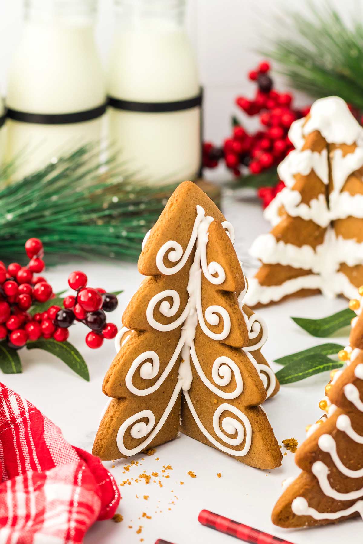 Three 3D Gingerbread Tree Cookies stand together, decorated with white icing; milk bottles and berries add a festive touch in the background.